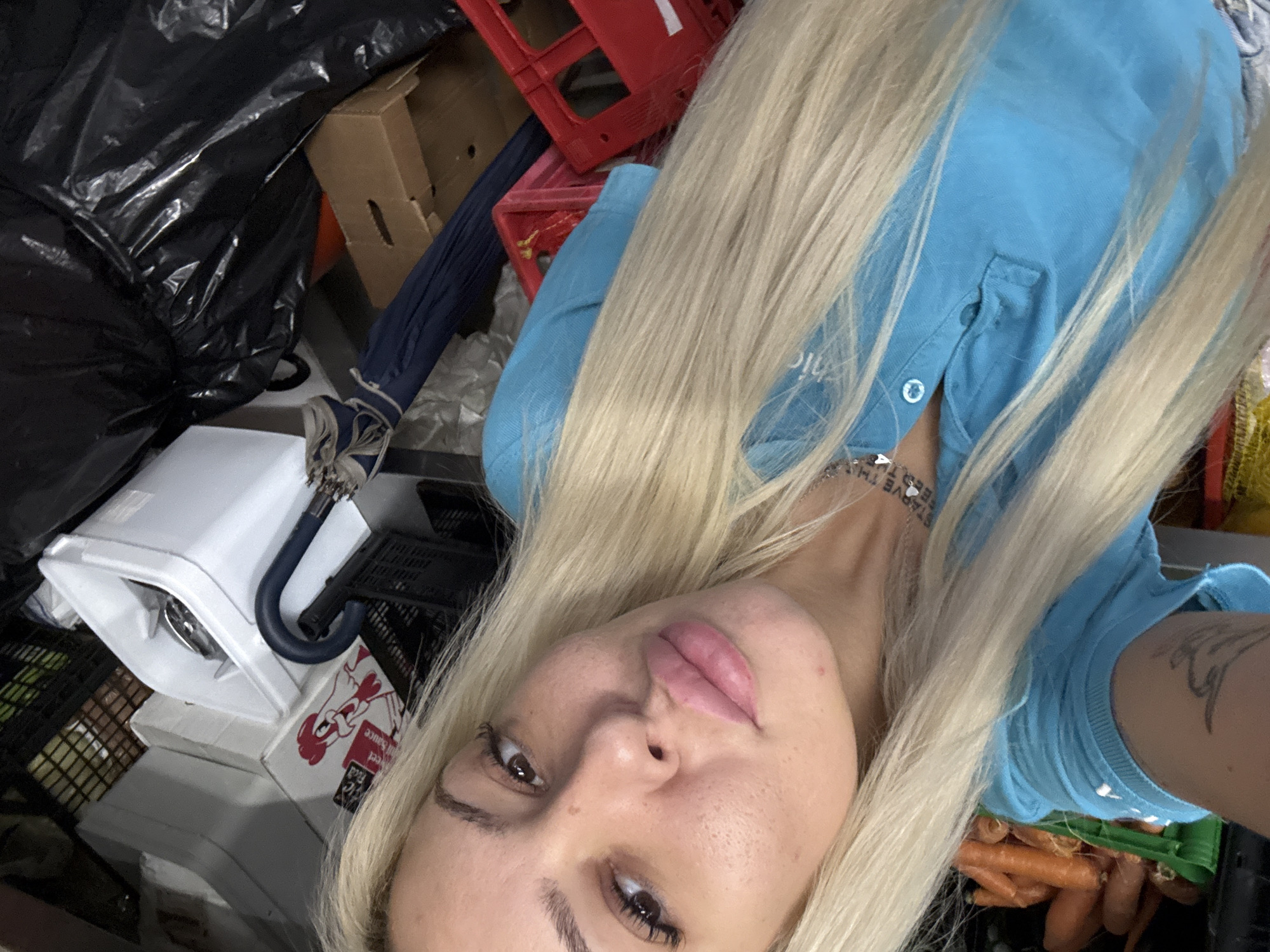 The image depicts a blonde woman in a blue shirt taking a selfie in what appears to be a storage area or backroom. She is surrounded by various items including black plastic bags, red and black plastic crates, a white appliance, an umbrella, boxes (one with "red sauce" printed on it), and produce such as carrots. It is not possible to determine the location in the city of Plovdiv, Bulgaria, without additional information.