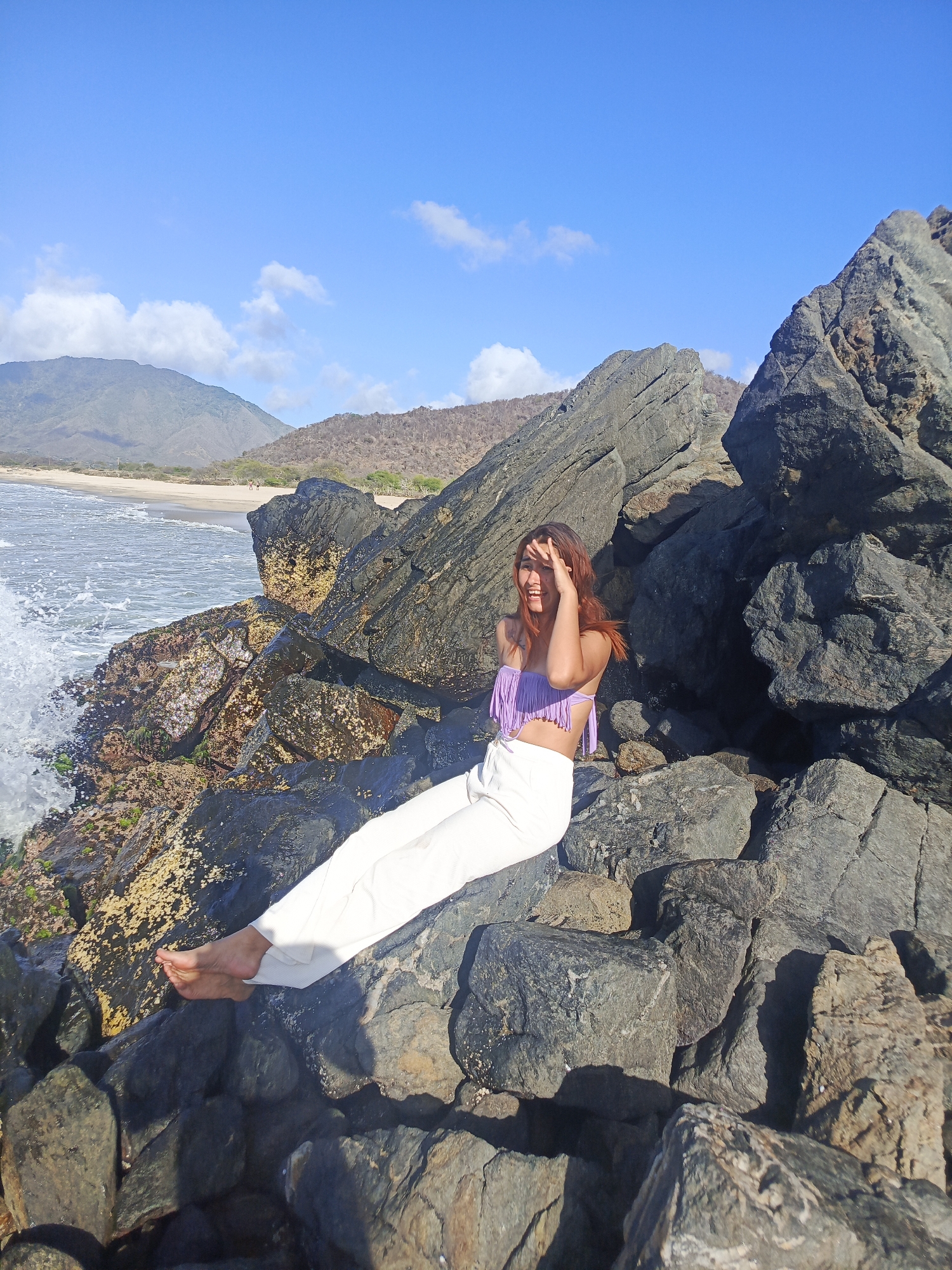 A young woman with long, reddish-brown hair sits on dark, rocky terrain near the ocean. She is wearing a light purple, fringed crop top and loose white pants. Her right hand is raised to shield her eyes from the sun as she smiles. The ocean, with waves crashing against the rocks, is to her left. In the background, a sandy beach meets dry, hilly terrain, with mountains visible in the distance under a clear blue sky with scattered white clouds. The location appears to be a coastal area in Pedro González, Venezuela.