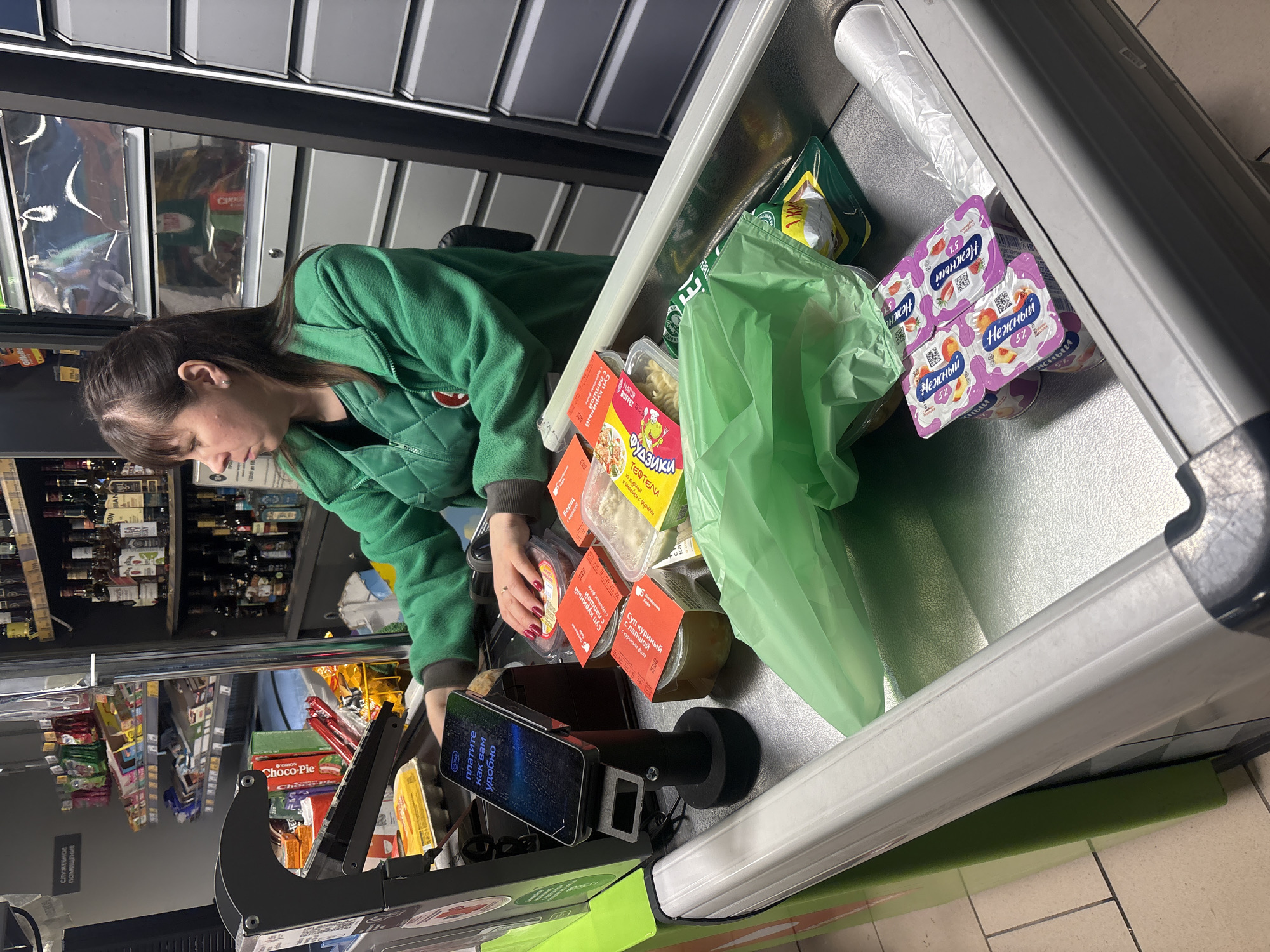 Here is a description of the image:
A medium shot captures a young woman in a green quilted jacket at a checkout counter in what appears to be a grocery store in Perm, Russia, based on the Cyrillic text on the packaging. She is reaching towards items on the conveyor belt, likely to arrange them for scanning. The conveyor belt holds various food products, including soups, pasta, yogurt labeled "Нежный" ("Nezhny"), and items in a green plastic bag. A payment terminal with a display screen showing payment options in Russian stands prominently in the foreground. Shelves stocked with various goods, including alcohol and snacks, are visible in the background. The overall lighting is bright, typical of a retail environment.
