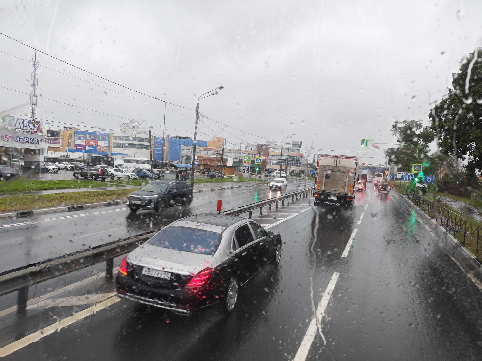 The image shows a rainy day on a multi-lane road in Lukhovitsy, Russia, with a view through a wet car windshield. The road is wet, with visible raindrops on the glass. Cars are driving on the road, including a black Mercedes-Benz sedan in the foreground on the right, a dark blue SUV on the left, and a large truck in the distance. Buildings and shops are visible on the left side of the road, including signs for "PER MARKET" and "KARI." Traffic lights are visible, with some showing green. Trees and a metal fence line the right side of the road. The sky is overcast.