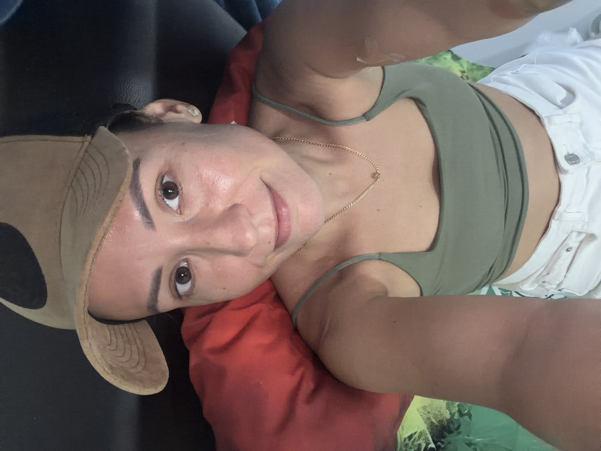 A woman lies on her back, propped up by a red pillow and a green and white patterned surface. She wears a tan baseball cap, a green tank top, and white shorts. Her head is tilted towards the camera, and she has a gold necklace on. The setting appears to be indoors, possibly a hotel room in Nha Trang, Vietnam, given the patterned surface which resembles a beach towel.
