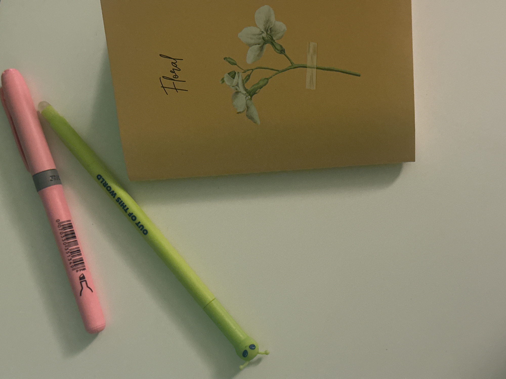 The image shows a close-up, high-angle shot of a white surface with two pens and a yellow notebook. The notebook is positioned in the lower right corner and features a delicate watercolor illustration of two white flowers with green stems, accompanied by the word "Floral" written in a script font. One pen, lime green with a small caterpillar-like figure at its tip, lies diagonally across the surface from the lower left to the upper right. The second pen, pink, is positioned above and to the left of the green pen, angled in the opposite direction. The surface appears to be a tabletop or desk. There is no visible indication of the city of Sofia, Bulgaria, or any human presence in the image.
