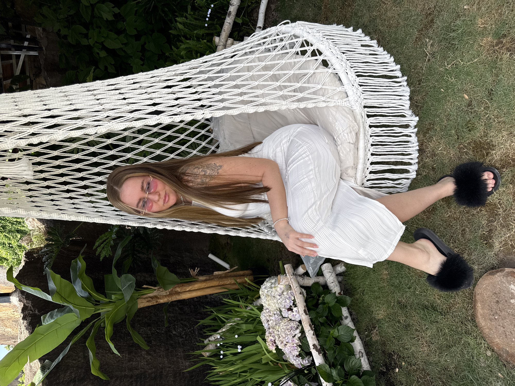 A woman is lying in a white hammock. She is wearing a white dress and black slippers with fur. The hammock is surrounded by greenery, including a banana tree and flowers. The hammock is supported by white birch branches. The location appears to be outdoors, possibly a garden or backyard. There is no indication of Keqiao District, China, in the image.