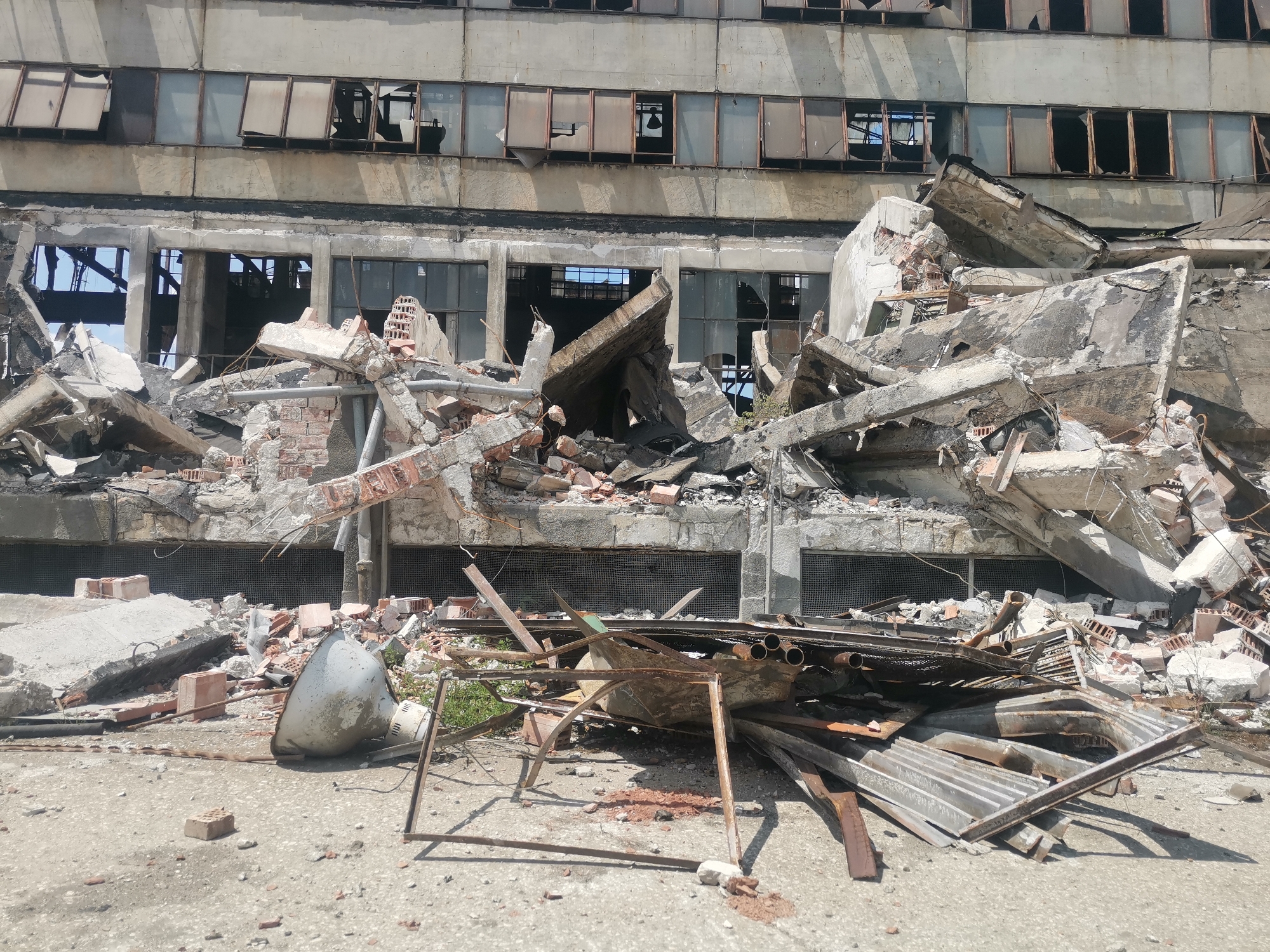 The image shows a destroyed building in Zhivkovo, Bulgaria. The structure is heavily damaged, with piles of concrete, bricks, and metal debris scattered in front of it. The building's facade is broken, with shattered windows and exposed interiors. There are no visible people in the image. The environment appears to be an urban area, judging from the paved ground and the presence of the building. The scene suggests a catastrophic event, such as an earthquake or war.