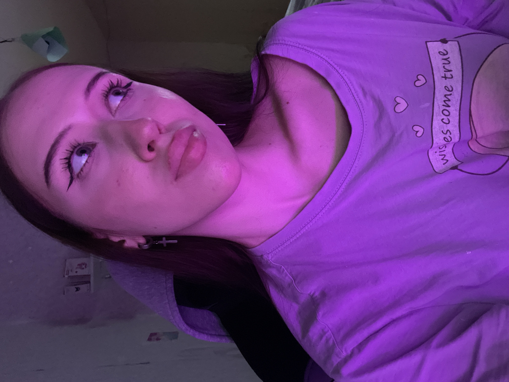 The media file is a vertical, close-up, self-portrait of a young person with long, dark hair and fair skin, bathed in purple light. The person is looking upwards and to their left. They have full lips, highlighted by lip gloss, and are wearing eye makeup with prominent winged eyeliner and mascara. A small stud is visible in their lower lip. A silver cross earring is worn in their left ear.
The person is wearing a light purple t-shirt with a graphic design on the chest. The design includes small hearts and a banner with the text "wishes come true" written in script.
The background is dimly lit and appears to be an indoor setting, possibly a bedroom. There are some indistinct objects visible in the background, including what looks like a shelf or table with items on it. The lighting creates a moody and intimate atmosphere. The time of day is not evident, but the indoor setting and artificial lighting suggest it could be evening or night. The location context of Votkinsk, Russia is not visually represented in the image. The overall emotion conveyed is introspective or perhaps wistful.