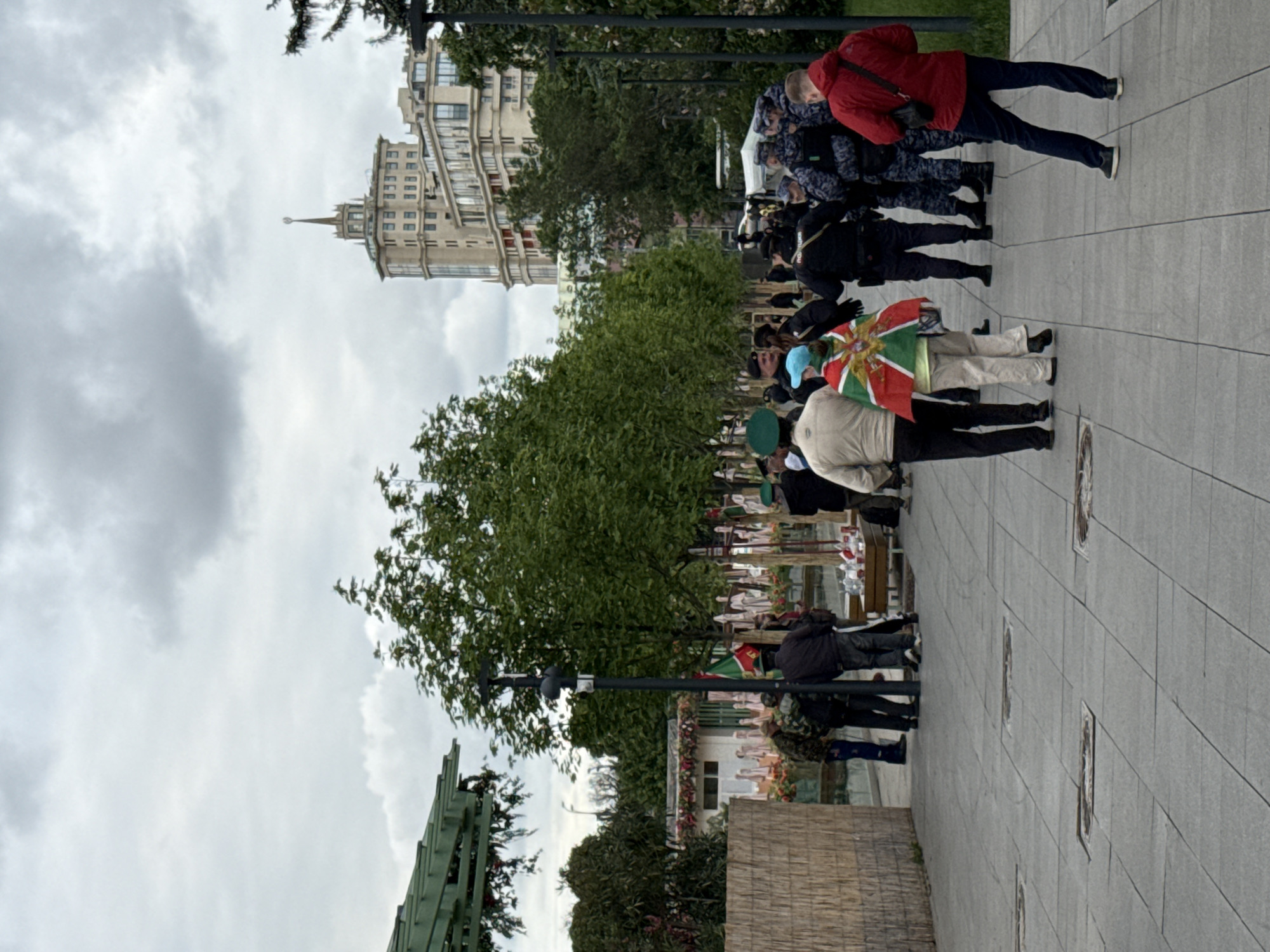 The image shows a group of people walking on a paved pathway. Some are wearing red jackets, while others are in police uniforms. One person is carrying a flag with a sun emblem. In the background, there is a tall building with a spire. The location appears to be in Moscow, Russia, given the architectural style of the building. Trees and lampposts are also visible, suggesting a park or public space.