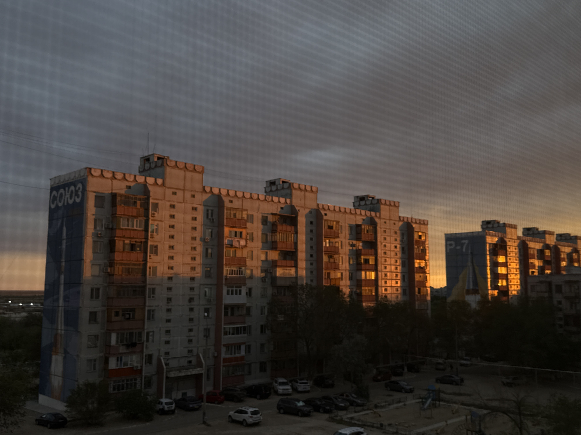 This is a photograph of apartment buildings in the city of Baikonur, Kazakhstan, during sunset. The sky is a mix of dark gray clouds and a warm orange glow from the setting sun. The buildings are multi-story residential structures with many windows. The building on the left has "СОЮЗ" (Soyuz) written in large blue letters on its side. The building in the center has a prominent "P-7" sign. There are cars parked in front of the buildings and trees scattered around the area. The scene appears to be a residential neighborhood.