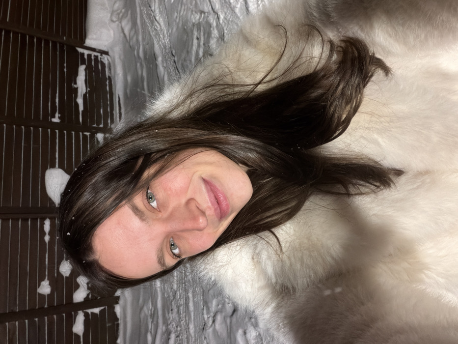 A woman with long, dark hair lies on her side, smiling at the camera. She has fair skin and green eyes. She is wearing a white fur coat or lying on a fur rug. Snowflakes dot her hair and the surrounding area. In the background, there is a wall or fence covered in snow. The scene appears to be outdoors in a snowy environment.