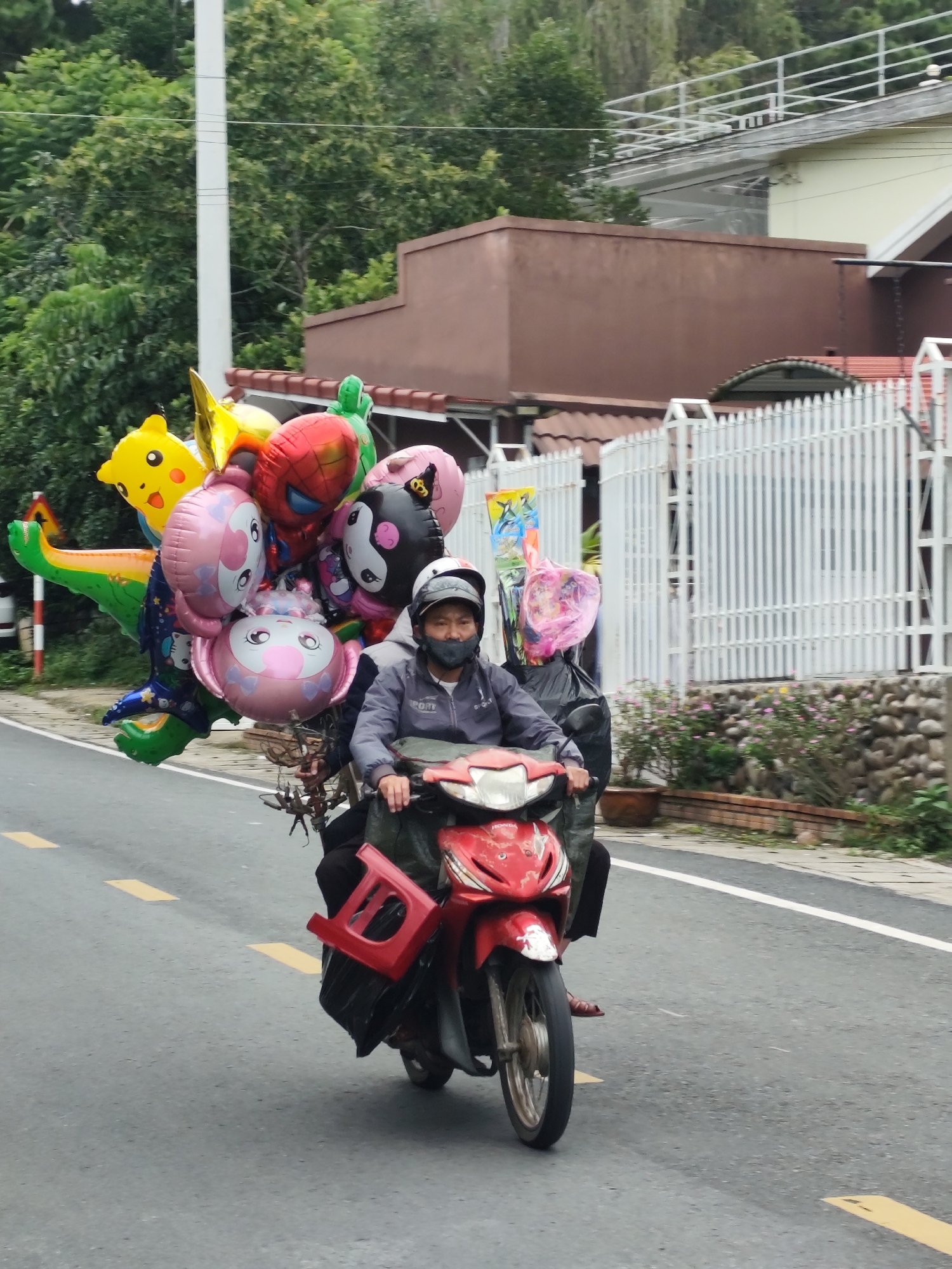 In Da Lat, Vietnam, a man on a red scooter navigates a paved road, his face obscured by a dark mask and helmet. He is transporting a large cluster of colorful balloons, including a green dinosaur, a yellow Pikachu, a red Spider-Man, and several pink cartoon characters. Behind him, another person is seated, also wearing a mask. The background features lush green trees and a white picket fence, suggesting a residential or semi-rural area. The road has yellow dashed lines indicating lanes. The overall scene captures a person going about their daily life, a common sight in the bustling streets of Vietnamese cities.