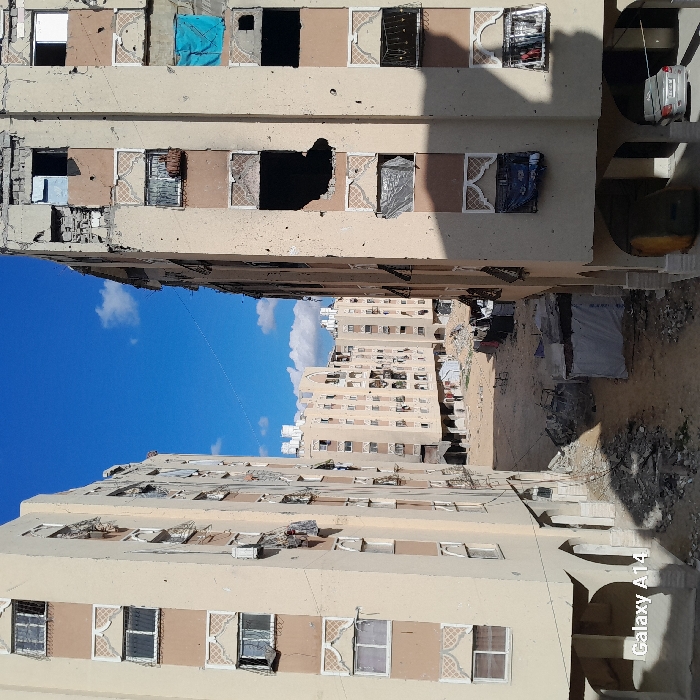 The scene depicts a devastated urban area in Khan Yunis, Palestinian Territory, characterized by heavily damaged multi-story residential buildings under a bright blue sky with scattered white clouds. Two light-beige apartment buildings frame the foreground, revealing a rubble-strewn, unpaved open ground area in the mid-ground, with more damaged buildings visible in the background.
The building on the left exhibits numerous broken windows, some covered with fabric or plastic, and several external air conditioning units. Its lower floors feature arched openings. The building on the right shows more extensive damage, including large, dark, irregular holes indicative of impacts on multiple floors, shattered window frames, and pockmarks across its facade. Specific windows are covered with a bright blue tarp, grey tarp, and dark fabric. Its ground level also includes arched openings, under one of which a light-colored sedan is parked, partially obscured by shadow.
The unpaved ground between the buildings is covered with piles of concrete rubble, sand, and debris. In the mid-ground, a small, makeshift shelter constructed from white tarpaulins is visible, alongside scattered personal belongings and what appears to be laundry hanging. A bicycle leans against debris near the center. In the background, multiple other multi-story residential structures, similarly appearing damaged, form a perimeter around the open area. No people or active interactions are visible.