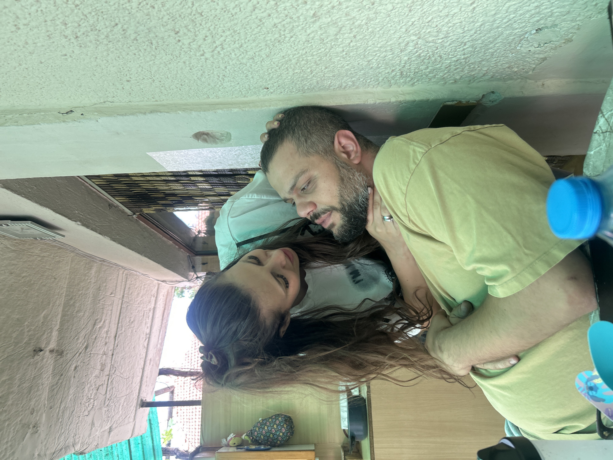 In Govedare, Bulgaria, a man with a beard and a woman with long, dark hair are shown in a tender moment. The man is leaning against a light green wall, his head tilted back. The woman is leaning over him, her hands gently touching his neck and head. Both are wearing casual clothing. The background suggests an outdoor or semi-outdoor setting, possibly a porch or patio, with a light-colored ceiling overhead and some wooden structures visible. The lighting is natural, suggesting daytime.