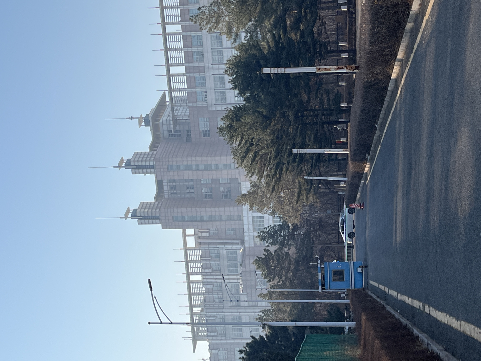 An eye-level, outdoor, medium shot reveals a street scene with buildings in the background, likely in Changchun City, China. The street, featuring a painted white line near its right edge, dominates the right side of the image. Adjacent to the street is a low hedge. Along the left side of the street, streetlights are visible. A blue automated license plate recognition (ALPR) camera enclosure is located on the left side of the road. A silver car is located further up the road next to orange traffic cones. High-rise buildings are partially obscured by trees and haze. The sky is a pale blue.
