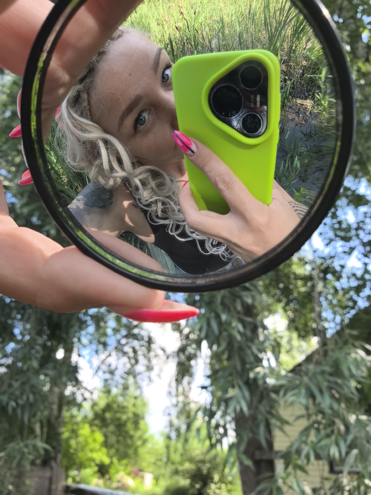 The image is a reflection in a circular mirror held by a person. The reflection shows a woman with blonde curly hair, blue eyes, and long, bright pink nails. She is holding a bright green smartphone with a triple-lens camera. The reflection also shows greenery and what appears to be a body of water in the background, suggesting an outdoor setting. The background of the image, outside the mirror's reflection, shows a blurry view of trees and foliage, with a hint of a building in the distance. The location is stated to be in the city of Noviy, Russia.