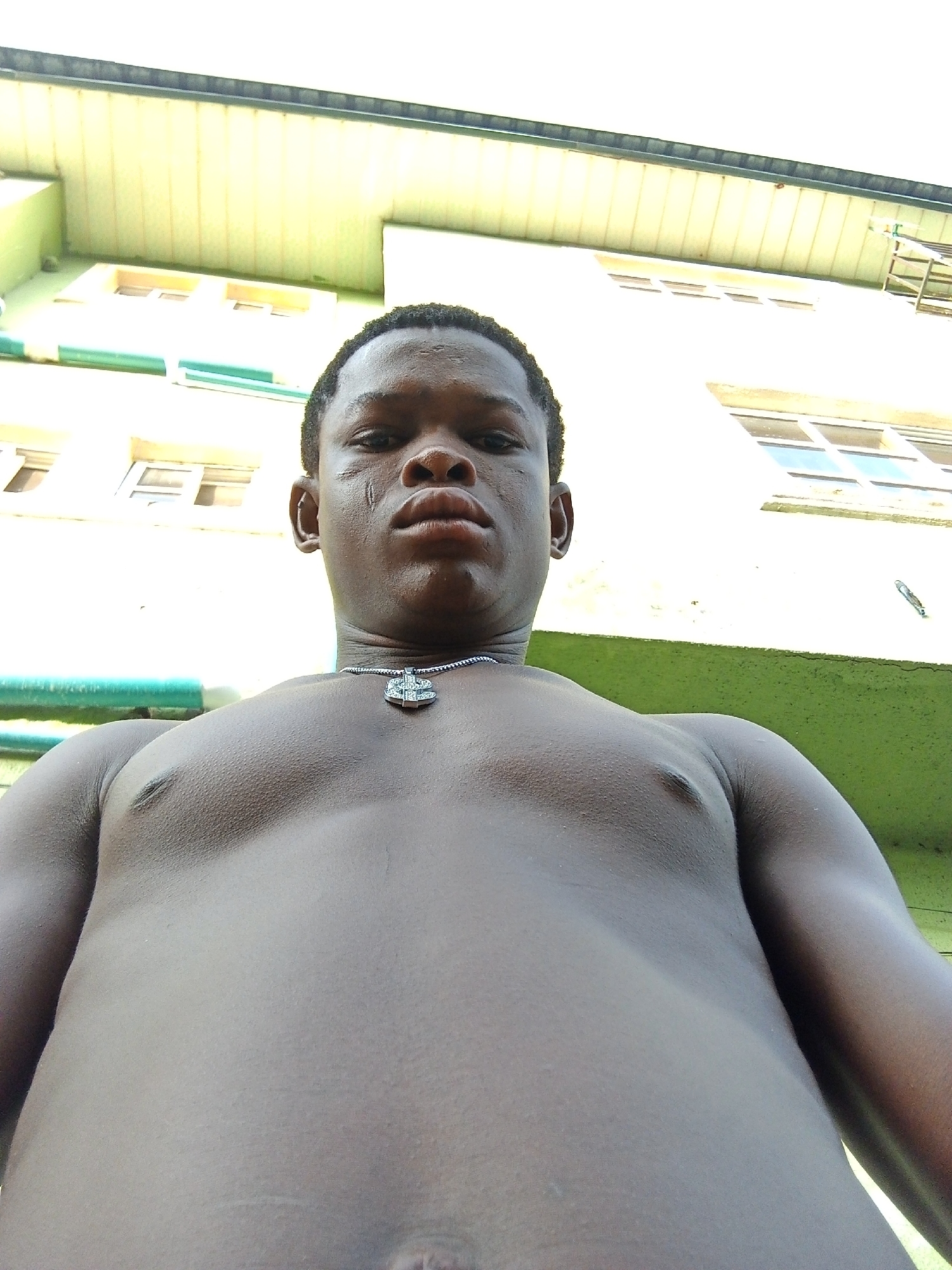 A close-up, low-angle shot depicts a shirtless Black male with dark skin, wearing a necklace with a dollar sign pendant. A visible scar marks his right cheek. The background shows a light green building with windows and possibly a ladder on the side. The location is plausibly within a city environment like Ado, Nigeria given the building style.