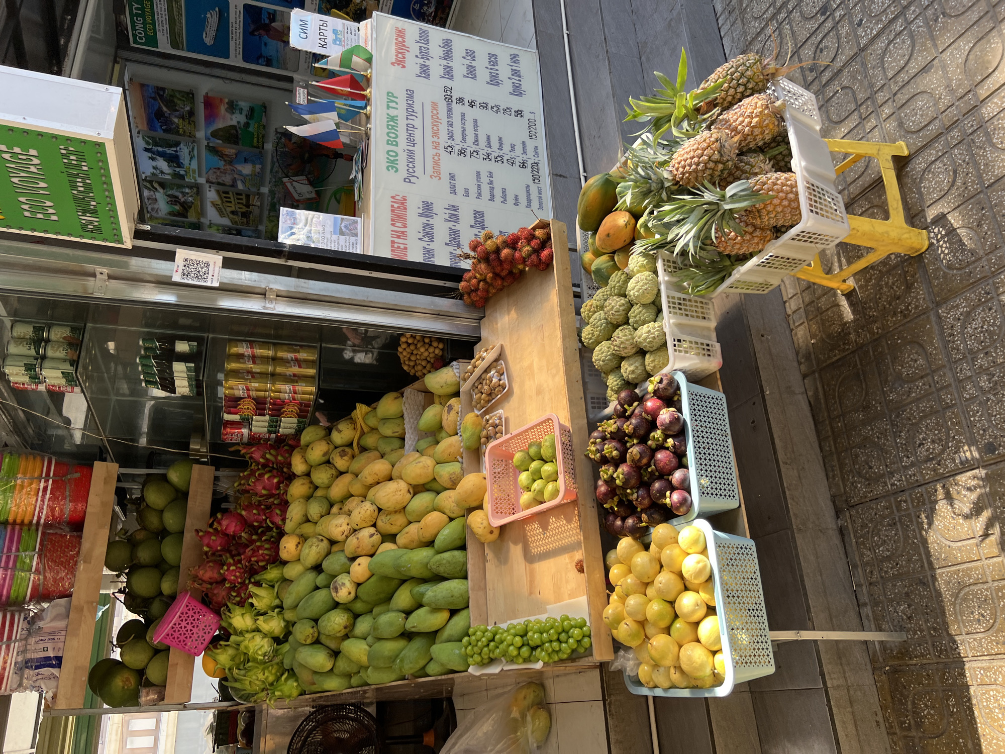 This is a street-level, eye-level, medium shot of a fruit stand in Nha Trang, Vietnam. The stand is laden with a variety of fresh fruits, including mangoes, dragon fruit, grapes, passion fruit, mangosteens, custard apples, papayas, and pineapples. The fruits are arranged in baskets and on wooden shelves. The stand appears to be part of a larger market or shop, as there are shelves with bottled drinks and what look like travel brochures visible in the background. The lighting suggests it is daytime, with sunlight casting shadows on the ground. There are no people visible in the frame. The ground is paved with patterned tiles.