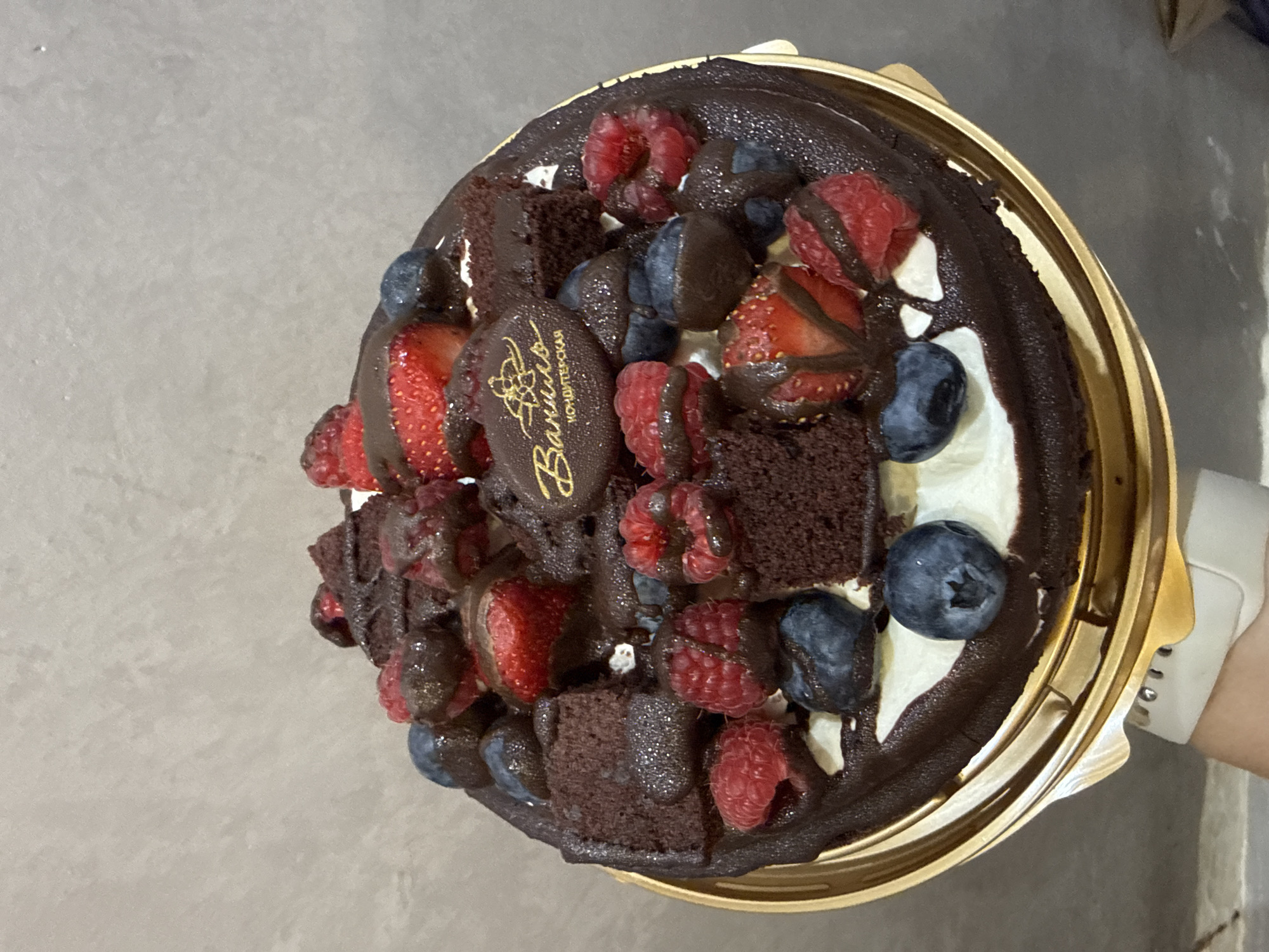 A high-angle shot shows a round chocolate cake in a golden-colored disposable container. The cake is topped with raspberries, blueberries, cut strawberries, and chocolate brownie pieces, all drizzled with dark chocolate. A chocolate plaque with the words "Ваниль" and "Кондитерская" in gold lettering is placed on top. Patches of white cream are visible between the fruit and brownie toppings. A hand wearing a white wrist-worn device can be seen holding the container. The background appears to be a neutral-colored surface. The city of Sochi, Russia is not discernible from the image.