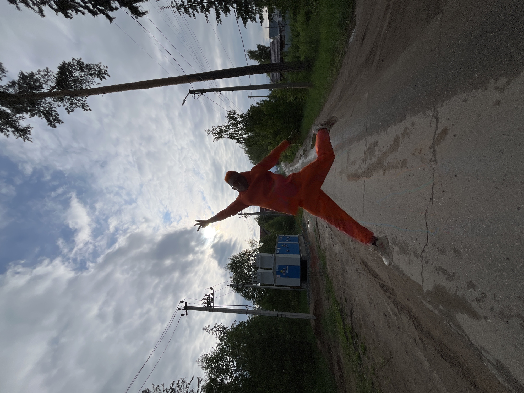 The image shows a person in an orange jumpsuit appearing to jump or strike a pose on a wet asphalt road. The person is in mid-air with one arm outstretched towards the sky. A row of power lines and poles are visible alongside the road, with trees and greenery in the background. There is also a blue and gray electrical transformer visible on the right side of the frame, near the bottom. The location is the city of Chaikovsky, Russia. The sky is overcast with clouds. The image is rotated slightly.