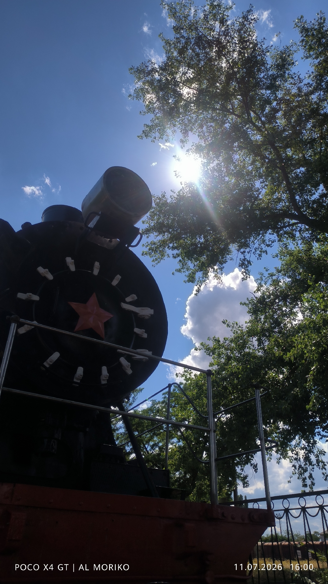 The image shows a close-up view of the front of a vintage steam locomotive under a bright blue sky with scattered clouds. The sun is shining intensely, creating lens flares and highlighting the leaves of a large tree on the right side of the frame. The locomotive is black with a prominent red star on its front. A metal railing runs in front of it. The bottom of the image displays text indicating "POCO X4 GT | AL MORIKO" and the date and time "11.07.2026 16:00". The scene appears to be an outdoor exhibition or historical site, possibly in the city of Bender, Moldova, given the text, though this specific location is not explicitly confirmed by visual elements within the image itself. There are no people or animals visible.