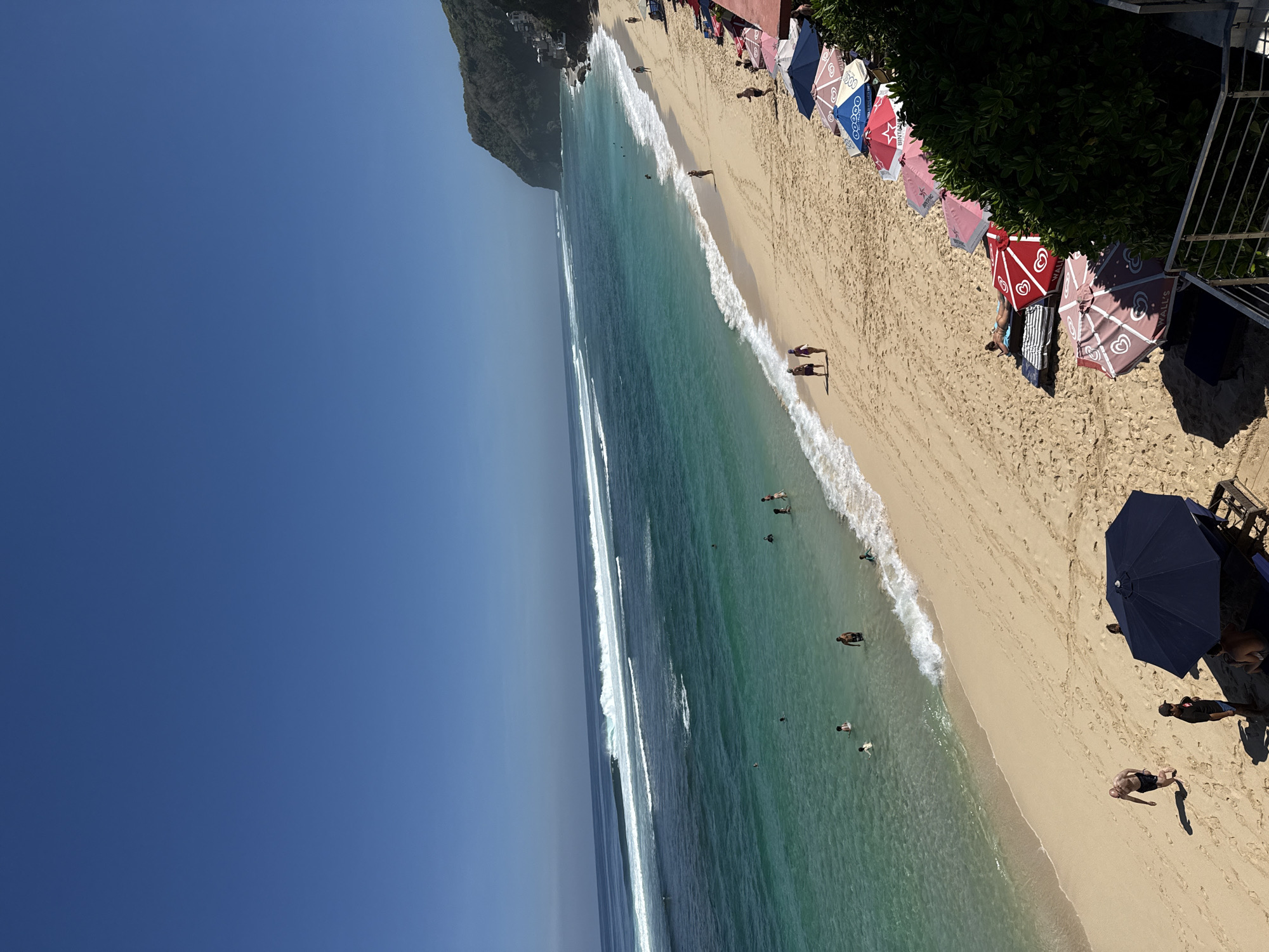 The image depicts a sunny day at a beach in Kuta Selatan, Indonesia, with a clear blue sky and calm turquoise waters. Numerous people are visible enjoying the beach and ocean. Several individuals are swimming or wading in the shallows, while others are walking along the sandy shore. In the foreground, a line of colorful beach umbrellas provides shade, with some people relaxing underneath them or sunbathing on lounge chairs. The beach itself is a wide expanse of golden sand, with gentle waves rolling onto the shore. In the distance, a rocky cliff face covered in lush greenery rises from the coastline, with buildings visible at the top. The scene conveys a relaxed and vibrant atmosphere, characteristic of a popular tourist destination.