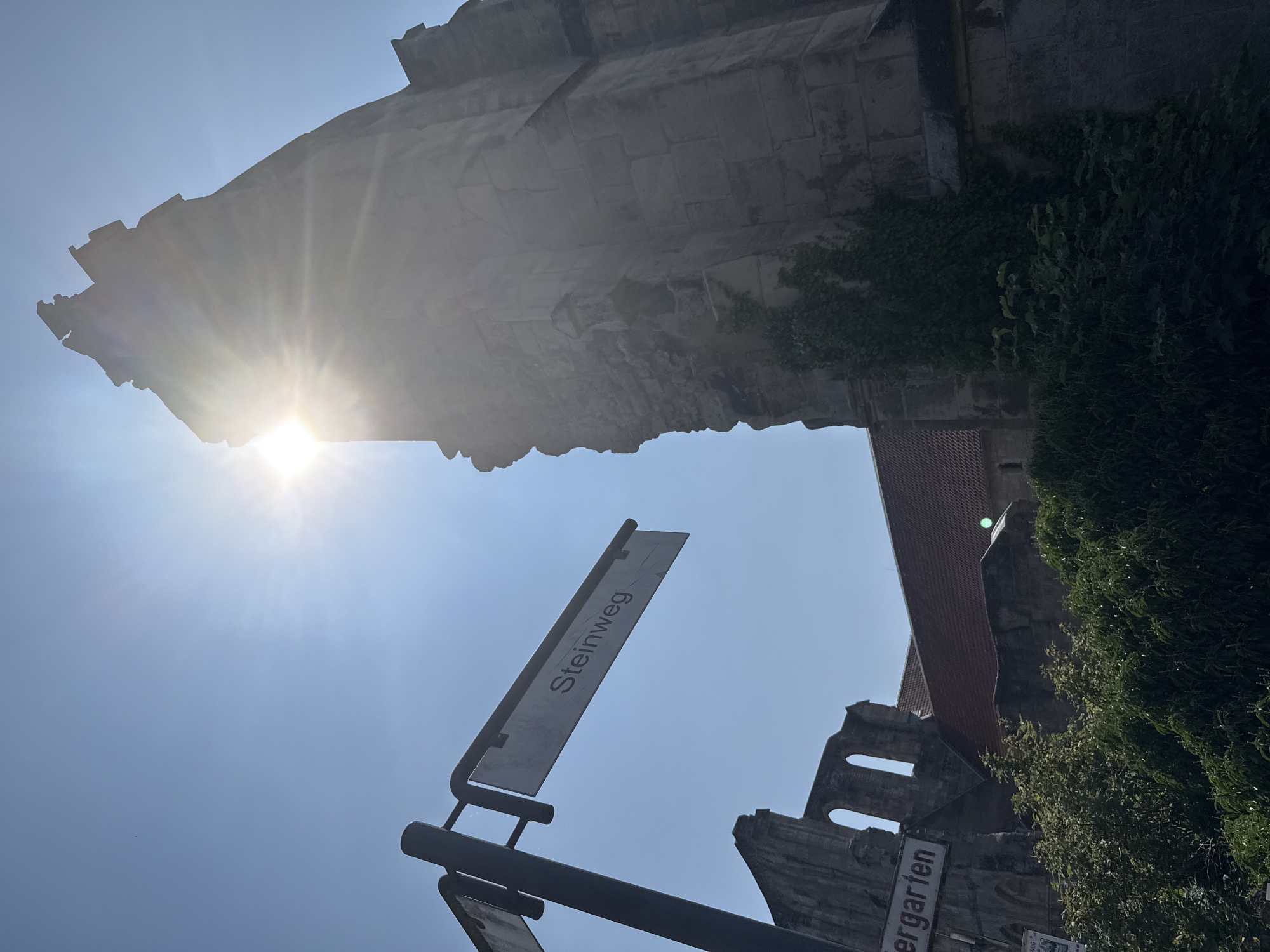 The image shows a street sign that reads "Steinweg". Behind the sign are the ruins of a large stone structure, possibly a church or abbey, with a red-tiled roof visible in the background. The sun is shining brightly, creating a lens flare effect and casting a glare on the scene. The ruins are partially overgrown with ivy and other greenery. The sky is clear and blue. The location is identified as Walkenried, Germany, likely referring to the Walkenried Abbey. There are no people visible in the image.
