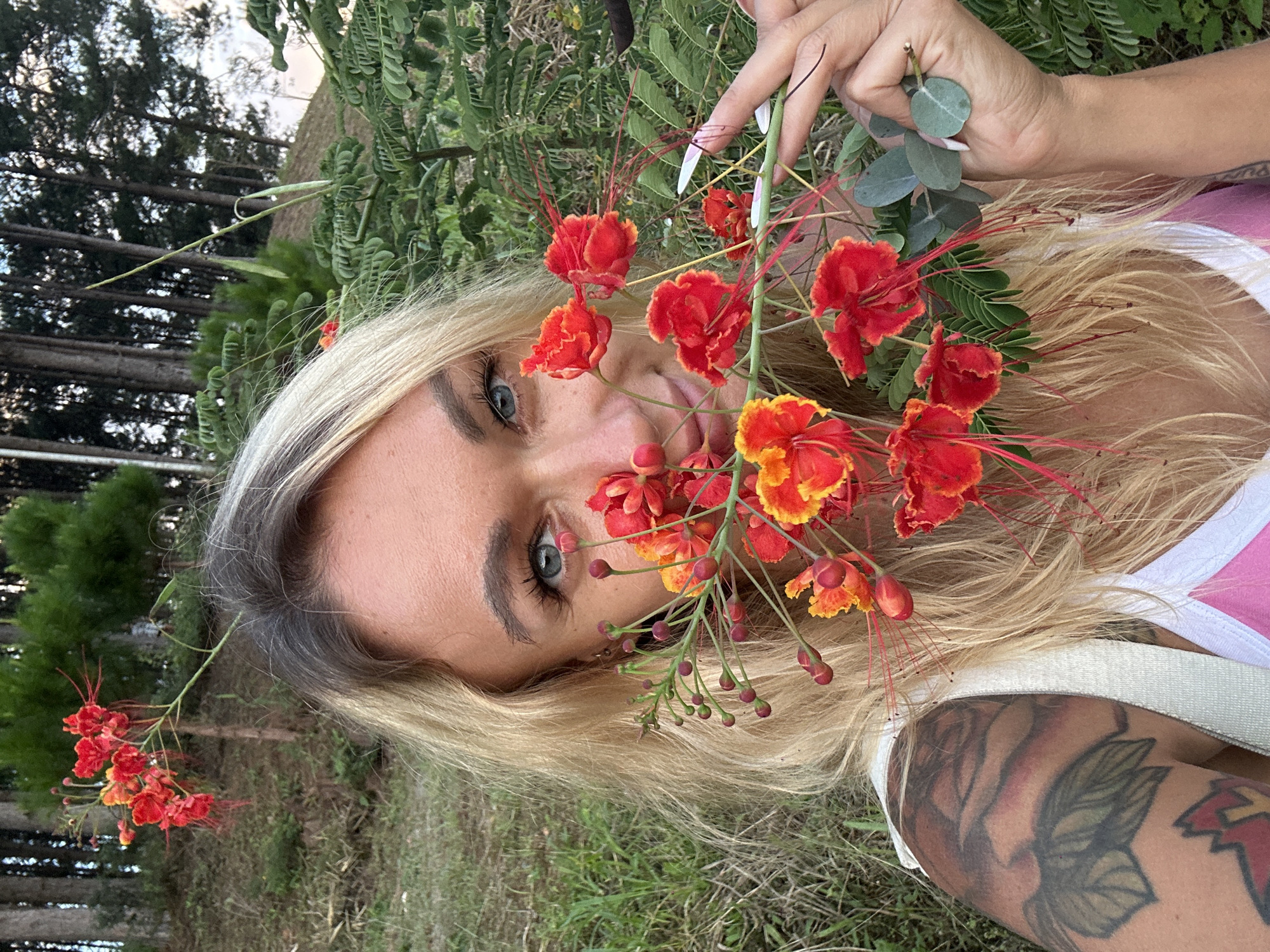 A blonde woman with blue eyes is taking a selfie outdoors, holding a branch with red and yellow flowers up to her face. She is wearing a white strap over a pink and white top. She has tattoos on her left shoulder and arm. The background consists of trees and grass, suggesting a natural or park-like setting. The location is specified as Thôn 3, Vietnam.