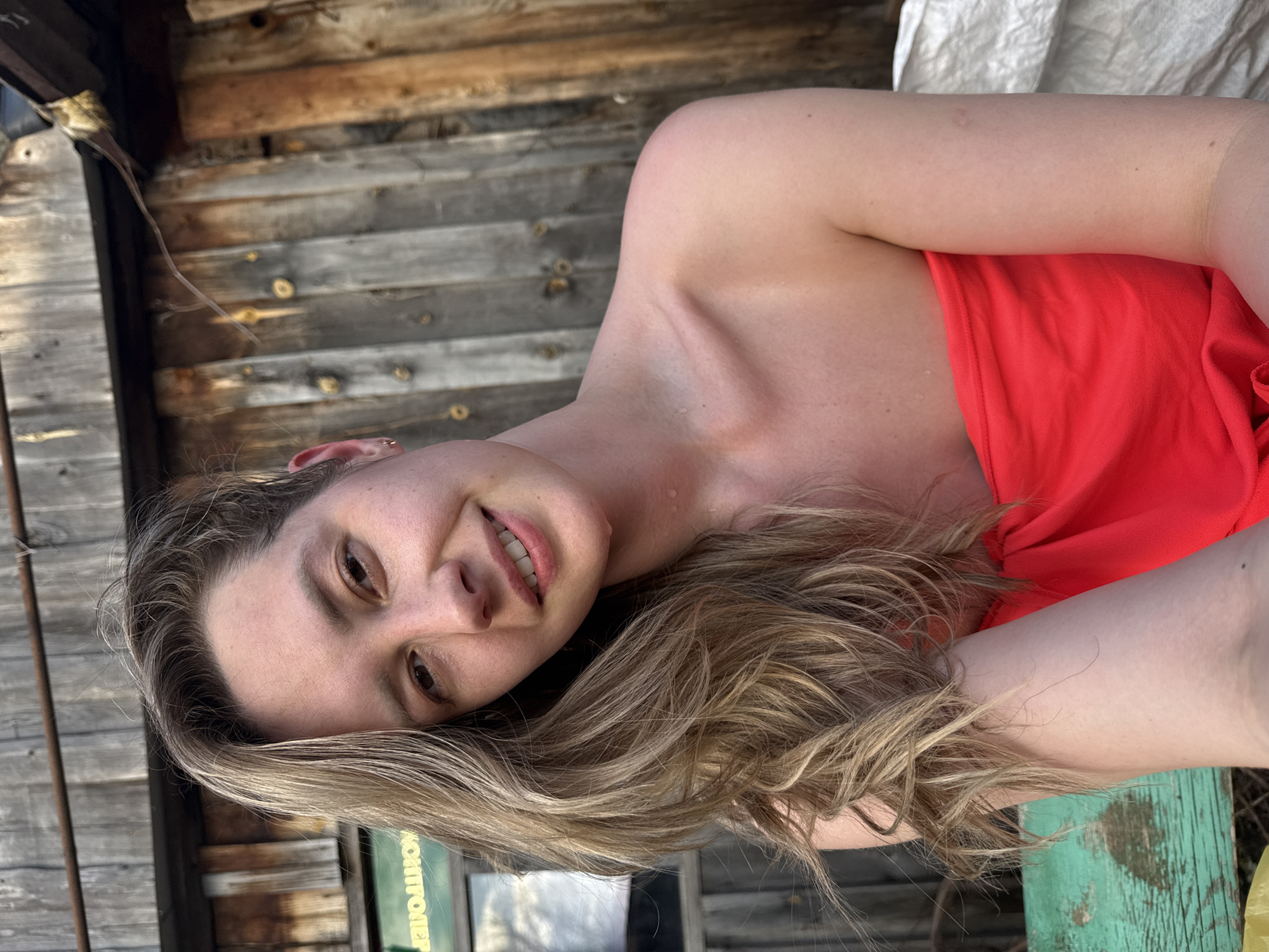 A woman with blonde hair, wearing a red garment that exposes her shoulder, is lying down in an outdoor setting. Behind her is a weathered wooden structure. A glimpse of the sky is visible through a window or opening in the background. Part of a word, possibly "мониторинг" (monitoring), which could indicate a utility or facility, is faintly visible. The precise location within the city of Chaikovsky, Russia is indeterminable from this image.