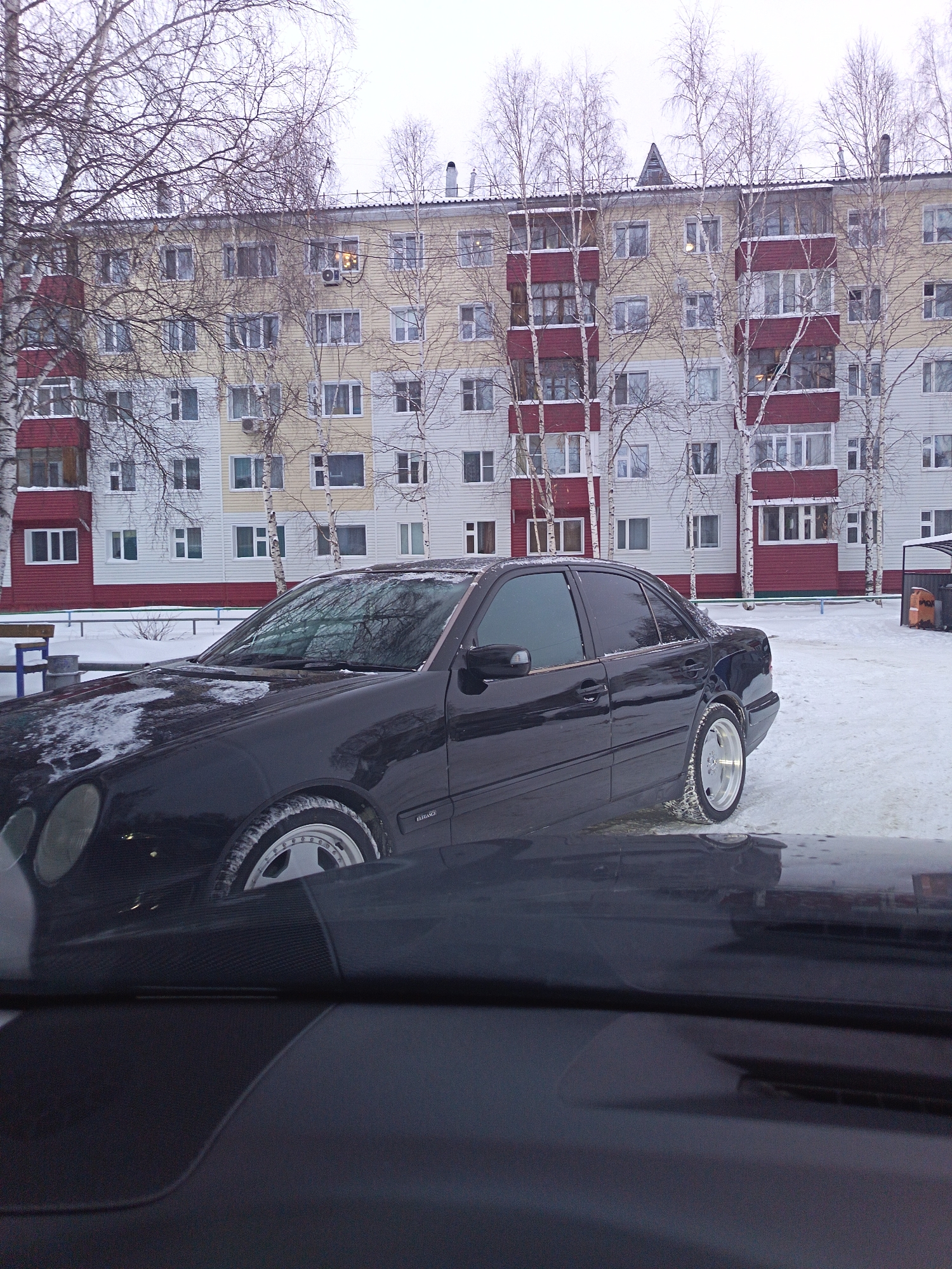 The image depicts a black Mercedes-Benz W210 parked in a snow-covered area in front of a multi-story residential building. The building has a facade with alternating bands of white, light yellow, and red-brown. Bare birch trees stand between the car and the building. The photo appears to be taken from inside another vehicle, with a portion of the dashboard visible in the foreground. The car is in Nizhnevartovsk, Russia. The ground is covered in snow, and the sky is overcast. A bench and some trash containers are visible in the background. The text "Elegance" is readable on the side of the car.