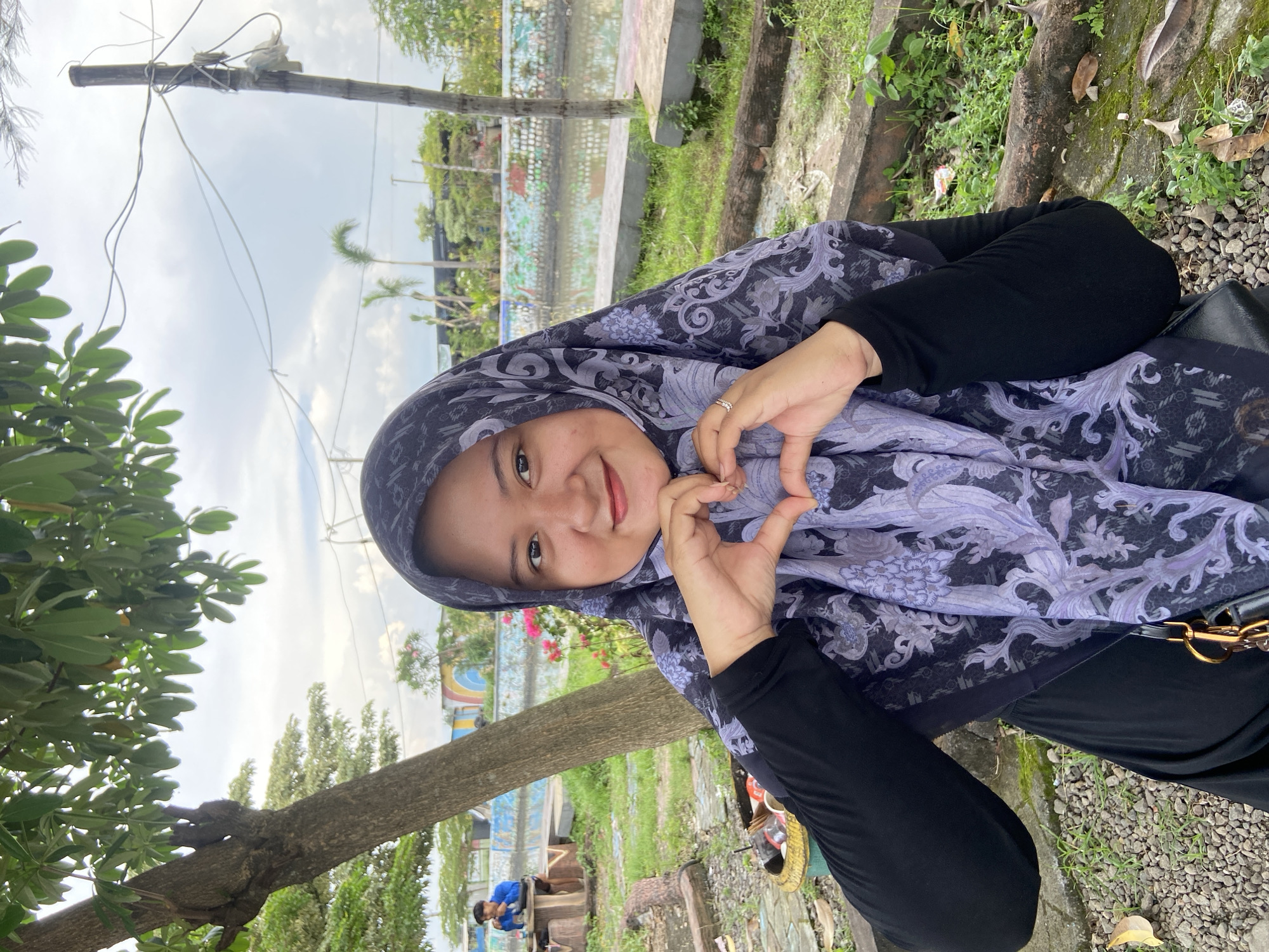 A woman wearing a hijab with a floral pattern is outdoors, possibly in Kalitengah, Indonesia. She is making a heart shape with her hands and smiling at the camera. She is wearing a black top. The background includes a body of water with a low wall. There are trees, grass, and pebbles visible. In the background, a man in blue is sitting on a wooden bench. Overhead is a utility pole and wires.