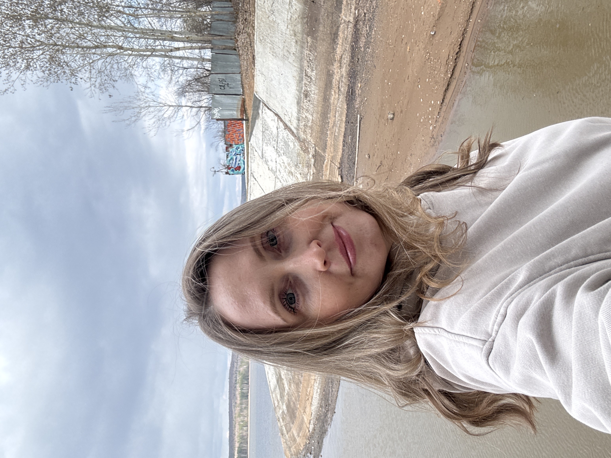 A young woman with long, light brown hair and blue eyes, wearing a light-colored (possibly beige or light grey) hoodie, takes a selfie outdoors. She is looking directly at the camera with a neutral to slight smile, her head tilted.
The background reveals a desaturated, industrial or semi-rural scene. To her left, a wide body of water, likely a river or large lake, stretches into the distance under an overcast sky. A barren, sandy, or muddy bank separates the water from a concrete structure, possibly a retaining wall or embankment, running horizontally across the middle ground.
Behind the concrete structure, a corrugated metal fence, covered in colorful graffiti, is visible. One section of the fence clearly shows the Cyrillic letters "СЕРБ" (SERB). Beyond the fence, a line of deciduous trees, bare of leaves, stands against the cloudy sky, suggesting late autumn, winter, or early spring. The overall light indicates daytime under an overcast sky.
The scene is set in Chaikovsky, Russia.