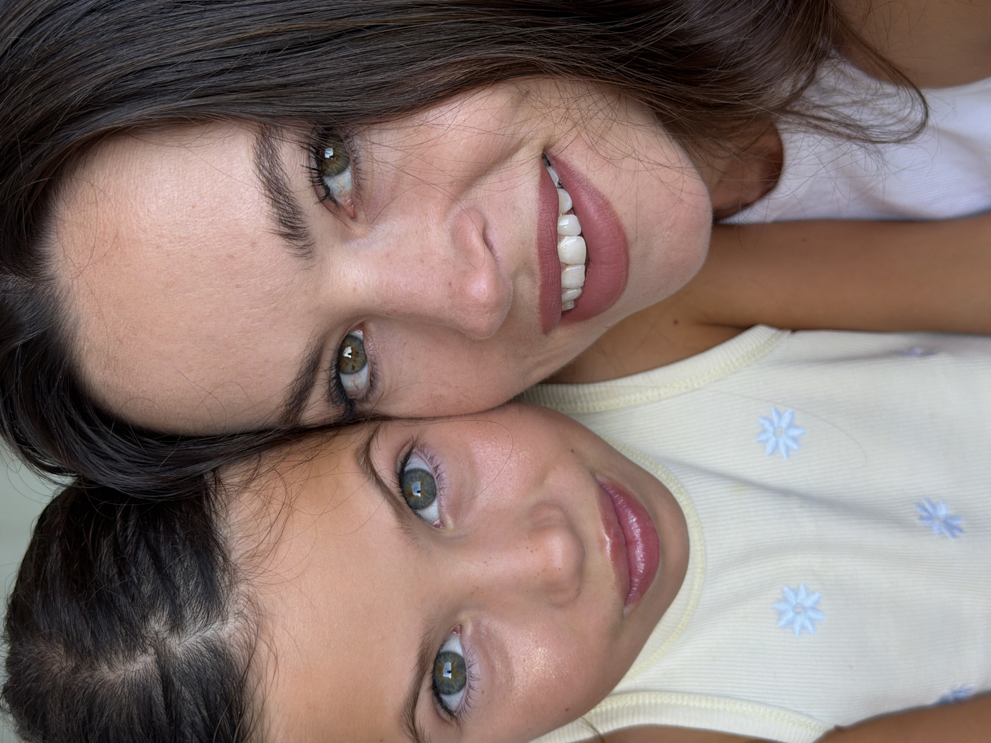 A close-up, head-and-shoulders shot features two females. The female on the left is a child with dark brown hair styled in a braid, her face turned slightly towards the viewer, with green eyes and a soft smile. She is wearing a light yellow ribbed tank top with blue floral embroidery. The female on the right is an adult with dark brown hair, her face also turned towards the viewer, with green eyes and a wider smile, revealing her teeth. She is wearing a white top. Their faces are close together, with the adult's head positioned slightly behind and to the right of the child's. The lighting is soft and natural, suggesting an indoor setting. There is no indication of the city of Salou, Spain, in the image.