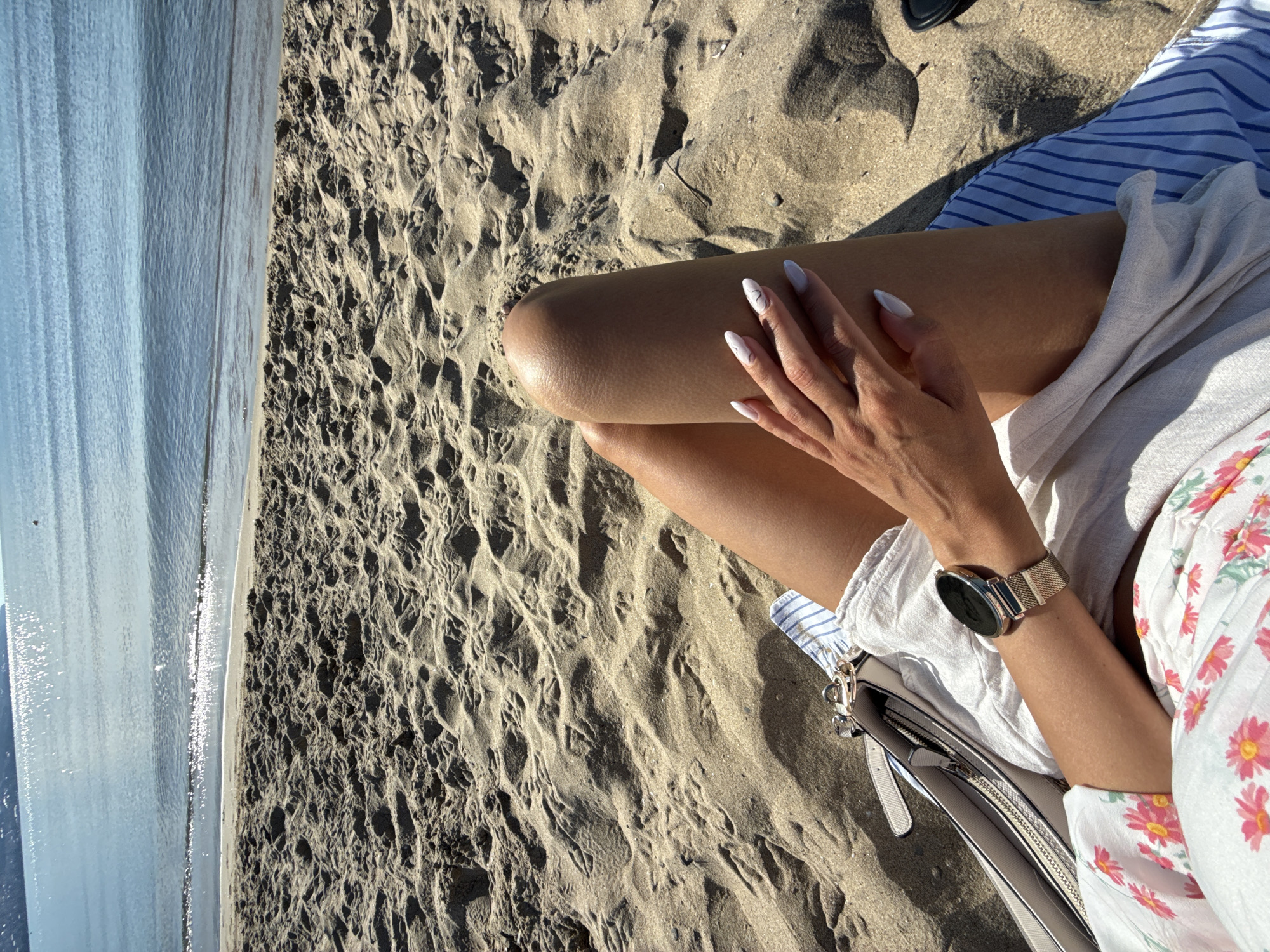 In Nesebar, Bulgaria, a woman is relaxing on a sandy beach with the Black Sea in the background. She is wearing a white top with a floral print, white shorts, and blue and white striped fabric. A silver watch is on her left wrist. A beige handbag is next to her. Her hand rests on her thigh, showcasing her white nail polish. The sand is textured with footprints, and the water is calm with sunlight reflecting off its surface.
