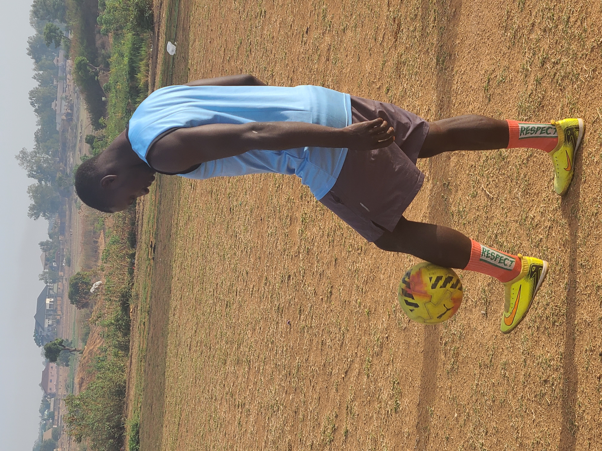 An outdoor, vertical shot captures a young man skillfully balancing a football on his right foot on a dry, earthy field in Barikin Jirga Summa, Nigeria. The man, with dark skin, wears a light blue sleeveless top and dark brown shorts. He is leaning forward, focused on the ball, with his head slightly down.
His feet are clad in vibrant orange socks that prominently display the word "RESPECT" in green lettering, paired with bright yellow and black football boots featuring a visible Nike logo. The football itself is yellow with red and black patterns. The ground is a mix of dry earth and sparse, light green grass, suggesting a hot, dry climate.
In the background, the scene transitions from the open field to a more rural or semi-urban landscape. Distant trees, scattered bushes, and simple residential buildings are visible under a hazy sky, indicating bright daylight, possibly midday or early afternoon. The overall impression is one of athletic practice or a posed shot showcasing football skills in a local setting.