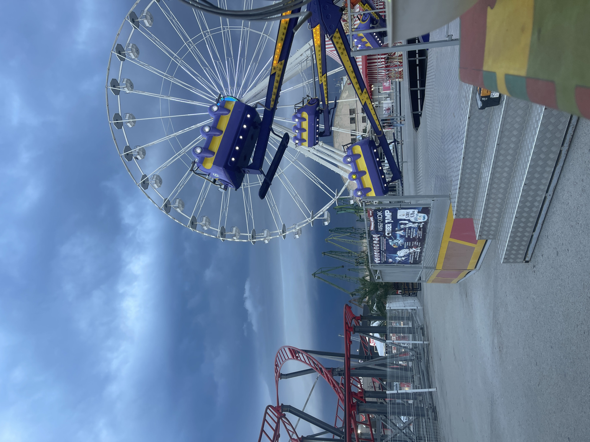 An amusement park is depicted under a dark, cloudy sky. In the foreground, a ride with multiple purple and yellow pods is visible. To the left, a red roller coaster track curves through the scene. In the background, a large Ferris wheel dominates the skyline. Several green industrial cranes are also present in the distance, suggesting a proximity to a port or industrial area. The ground is paved, and metal stairs lead up to a platform. A poster for "CYBER JUMP" is displayed on a stand near the foreground ride. The overall atmosphere is somewhat ominous due to the stormy weather. The location is the Varna amusement park in Varna, Bulgaria.