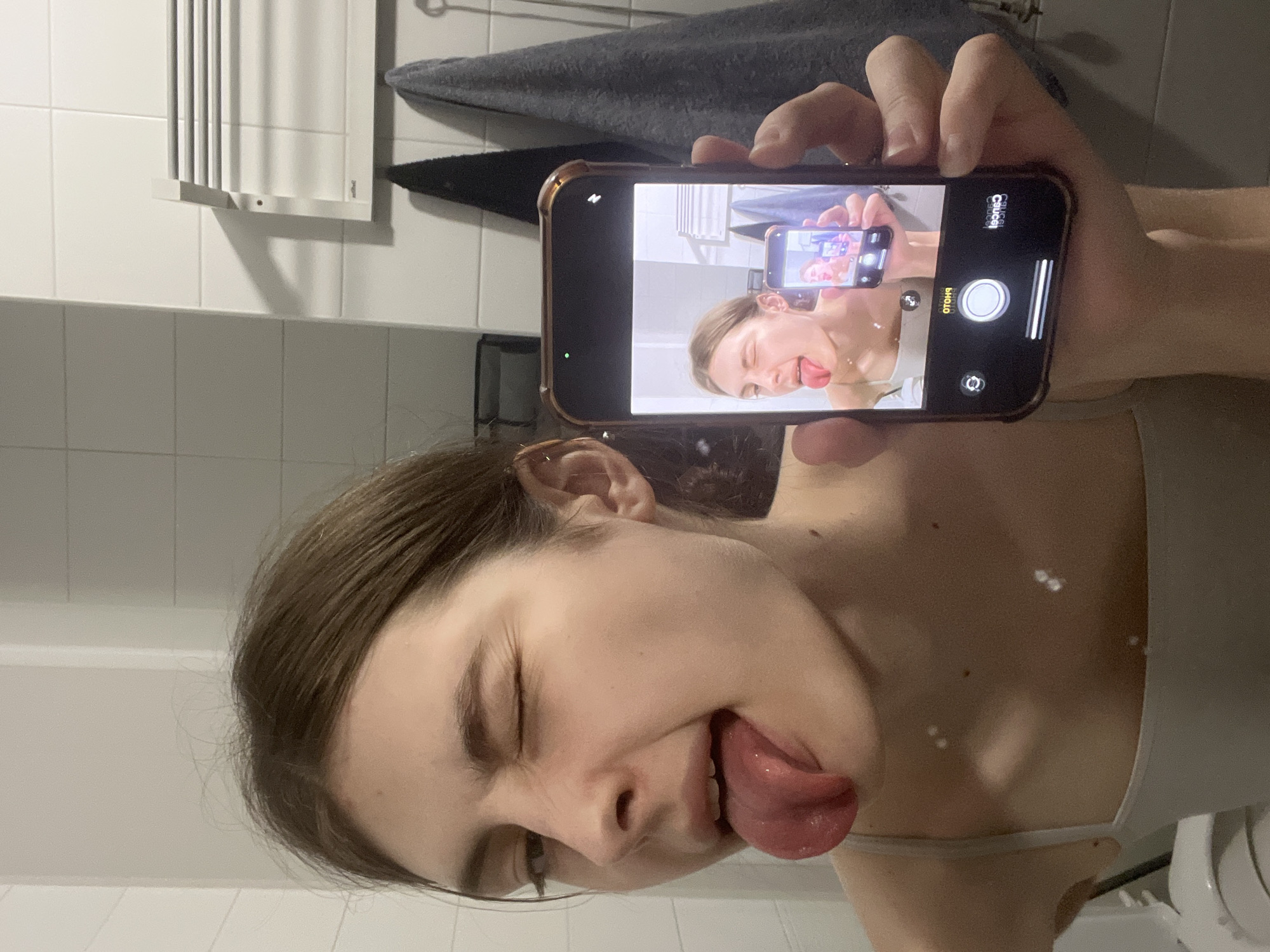 A woman is holding a smartphone in her right hand, taking a picture of herself in the mirror. The phone in her hand shows the image of herself sticking her tongue out. The background shows a bathroom with white tiled walls and a towel rack. The location is not identifiable as Saint Petersburg, Russia.