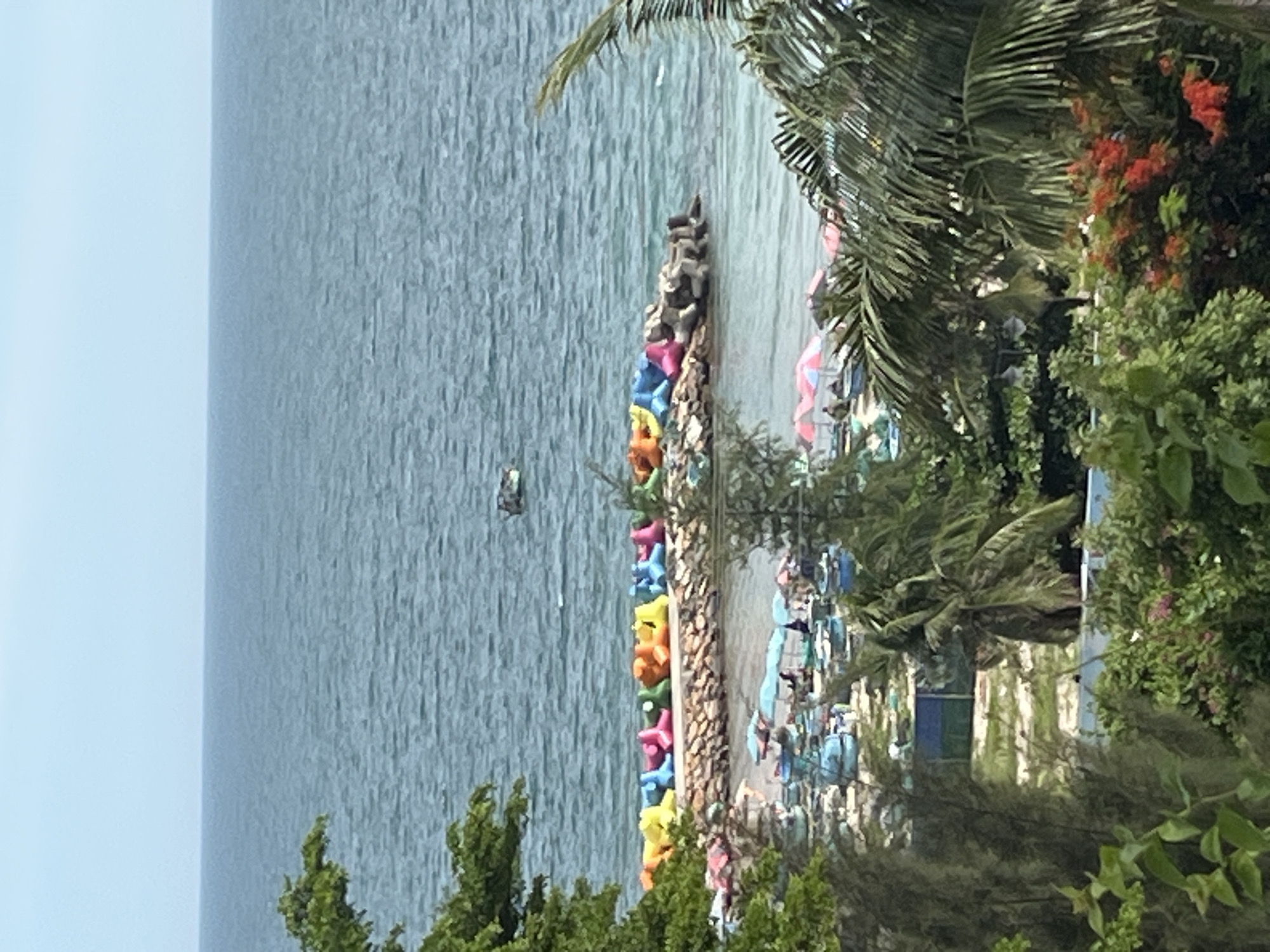 The image shows a coastal scene in Phu Thuy Ward, Vietnam. In the foreground, lush green foliage and flowering plants are visible. Behind this, a section of a beach is apparent with numerous blue fishing boats and several pink and red umbrellas providing shade. In the mid-ground, a stone breakwater separates the beach from the sea. Along the top of the breakwater, a row of colorful, inflatable beach toys are arranged, including yellow, orange, pink, and blue shapes. The sea is calm and extends to the horizon under a hazy blue sky. A small boat is visible in the distance on the water.