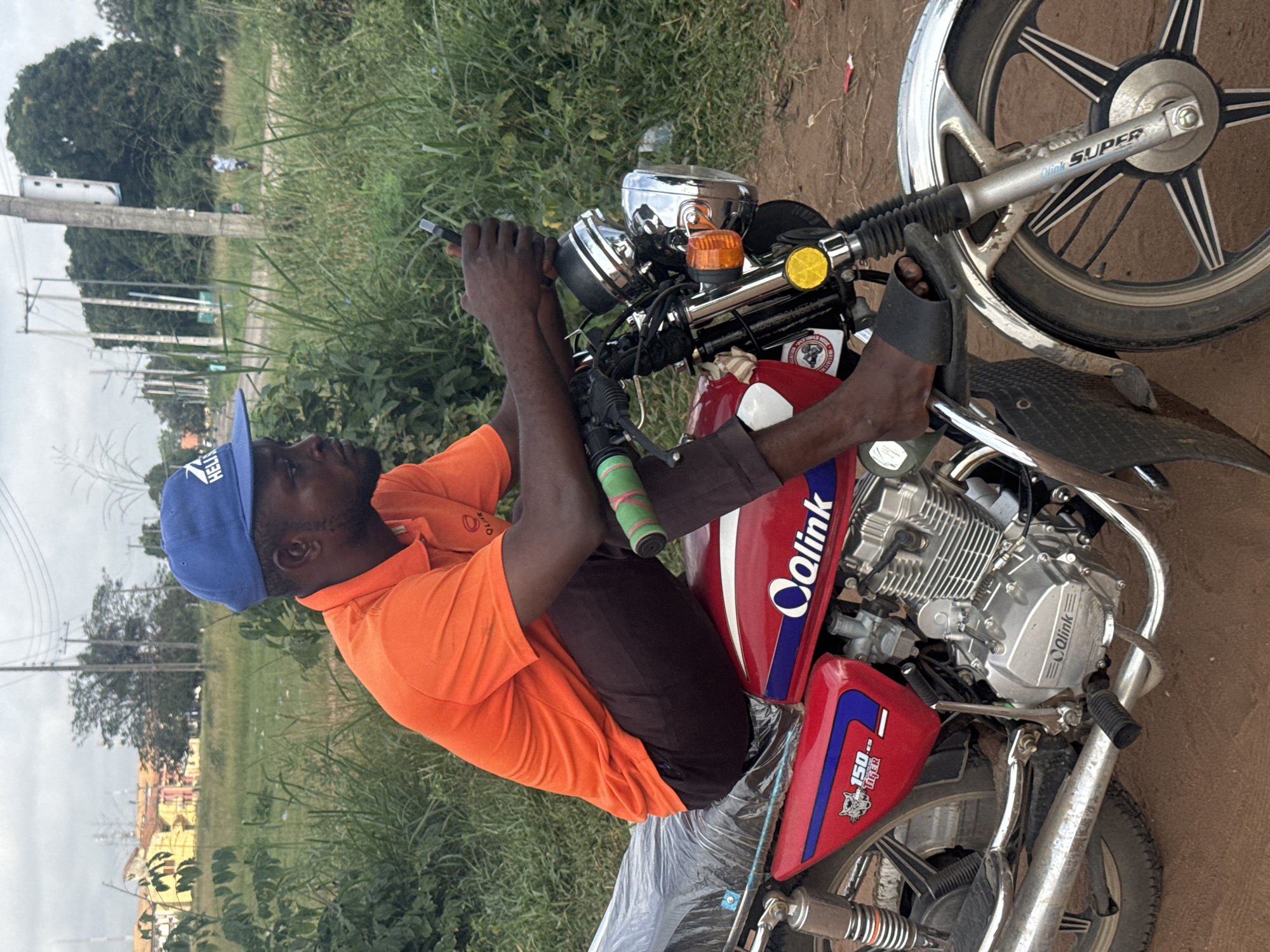 The image, taken in Ihiagwa, Nigeria, features a man seated on a red and blue Qalink brand motorcycle. The man wears an orange short-sleeved polo shirt, dark trousers, and a blue baseball cap with the partial word "HELL" visible. He is holding the handlebars with both hands and looks towards the left of the frame with a neutral expression, his feet resting in black sandals.
The motorcycle's fuel tank is red with blue accents and features the "Qalink" logo, along with "150 Tiger" text and an emblem of a roaring animal. The silver engine block is also embossed with "Qalink." Chrome components are visible around the headlight and other parts of the bike. The front suspension forks, labeled "SUPER" and "Qalink," hold a wheel with a distinctive multi-spoke design. An orange turn signal and a yellow circular reflector are present near the handlebars. Part of the motorcycle's seat or rear fender is covered with a clear plastic sheet.
The setting is outdoors, likely a roadside or dirt path, characterized by light brown earth. Abundant green grass and shrubs flank the path, extending into the background. Under an overcast sky, tall trees and utility poles with power lines are visible in the distance, alongside some indistinct distant buildings. The scene appears to be taking place during the daytime.