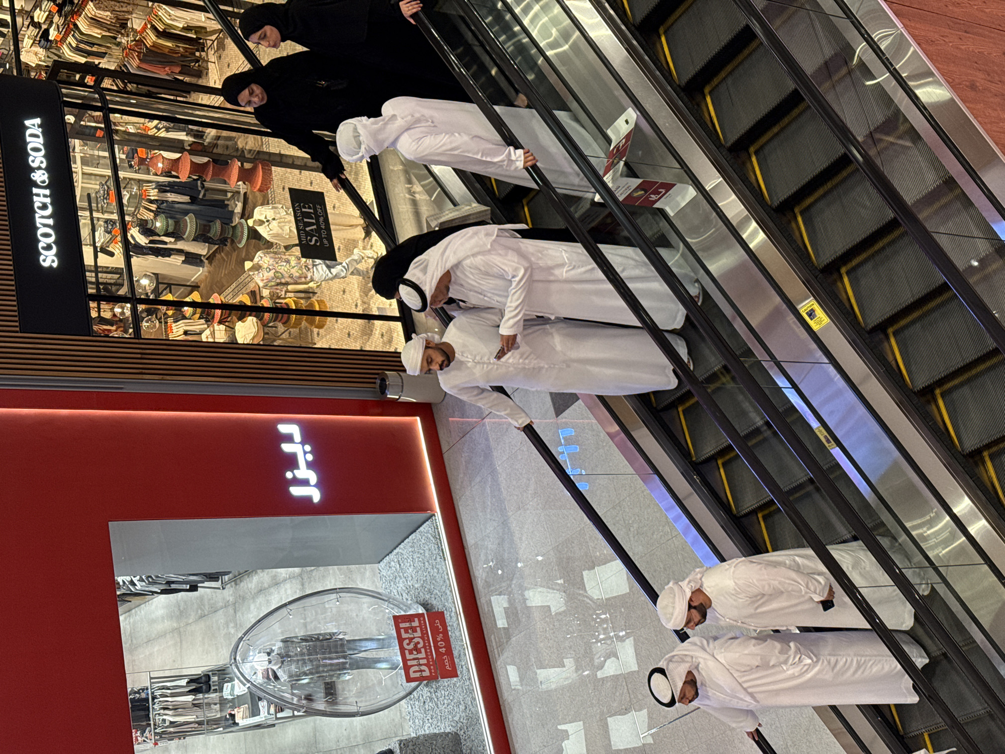 The image shows an escalator in what appears to be a shopping mall in Dubai, United Arab Emirates. Several individuals are riding the escalator. Two women dressed in black abayas and hijab are at the top. Below them is a person in a white thawb and ghutra. Further down are several men also dressed in traditional white clothing including thawbs and ghutras, suggesting they are local Arab men.
The escalator runs past storefronts. One is for the brand "Scotch & Soda" which has a sign advertising a "Mid Season Sale" with up to 40% off. Another storefront visible is for "DIESEL". The store also has a sign stating "Up to 40% off".