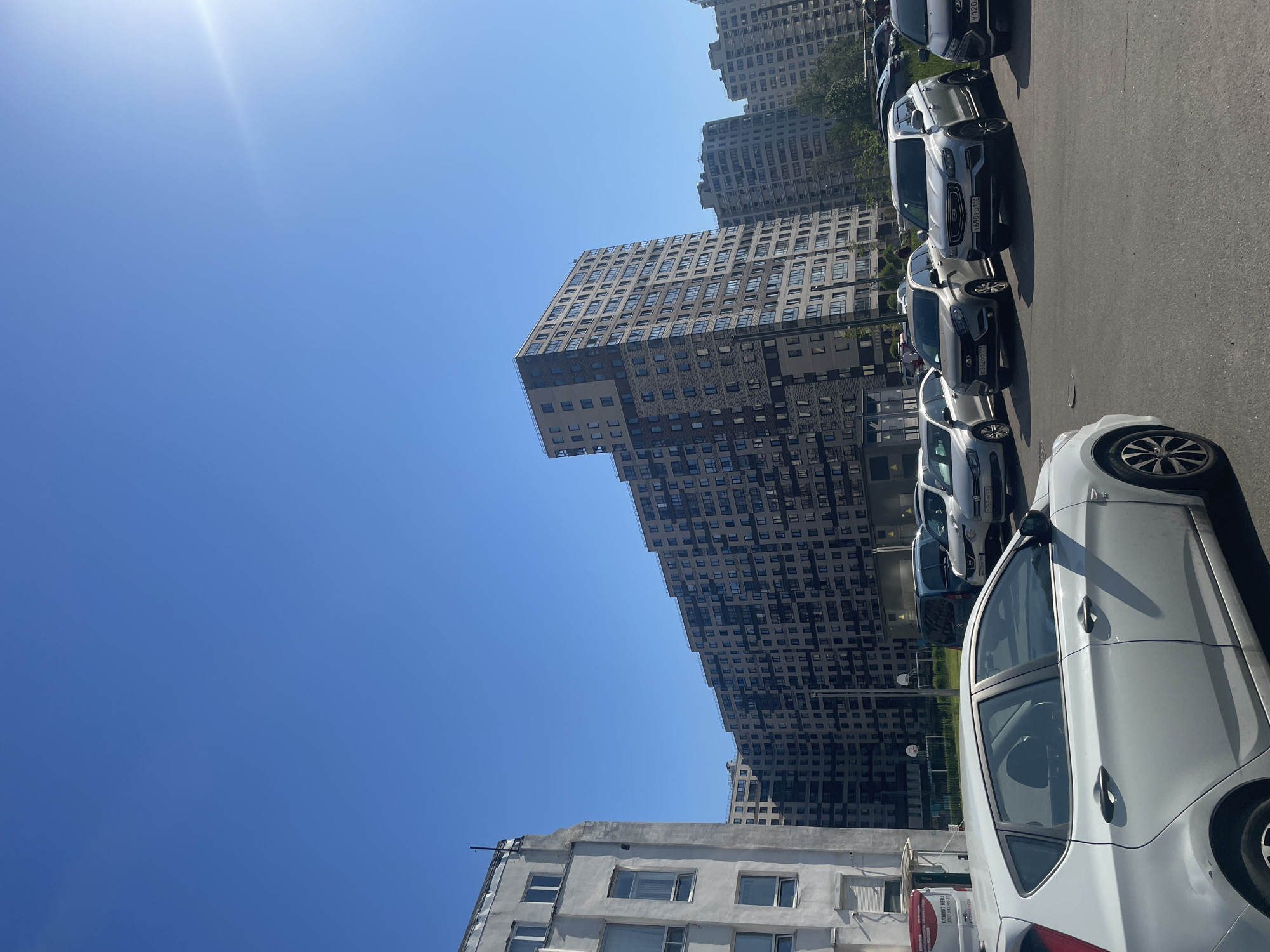 The image captures a bright, sunny daytime street scene in an urban area, likely in Russia as indicated by text and license plates. The photograph is oriented vertically, rotated 90 degrees clockwise from a standard landscape view.
In the background, several tall, modern residential buildings with numerous windows rise into a clear blue sky. One prominent, large building features a distinct, stepped or staggered design with varying facade patterns. Closer to the foreground, a lower, older-looking white building is partially visible on the left.
The foreground is dominated by a row of parked cars along a paved street or parking lot. A white five-door hatchback is prominently featured on the right side of the frame. Behind it, other vehicles, including SUVs and sedans, are parked in a line.
The sky is a vivid, cloudless blue, suggesting fair weather. A bright sun flare is visible in the top-left corner, indicating the presence of direct, intense sunlight, likely during late morning or early afternoon. Shadows cast by the cars and buildings are sharp and well-defined.
Visible text on a sign or sticker in the lower left includes "КЛИМАТ НЕВА" (KLIMAT NEVA) and a phone number "(812) 642-40-20," where "812" is a Saint Petersburg area code. Russian license plates are visible on several cars, featuring codes such as "Р690ТО 198" and "Е332ОО 198," with "198" indicating the Moscow region. These details collectively point to a Russian location.