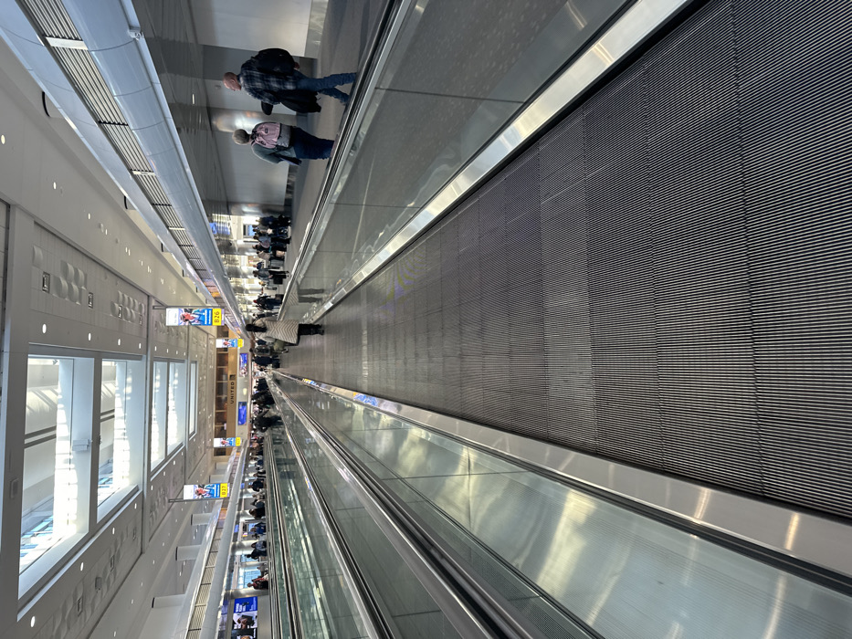 An interior shot captures a long moving walkway in an airport or similar public space. People are sparsely spread along the walkway, with a small cluster at the far end. Large windows are visible on one side. Signage hangs from the ceiling advertising gates. The walkway's surface is lined with horizontal grooves. The lighting is bright, and the overall atmosphere is sterile.