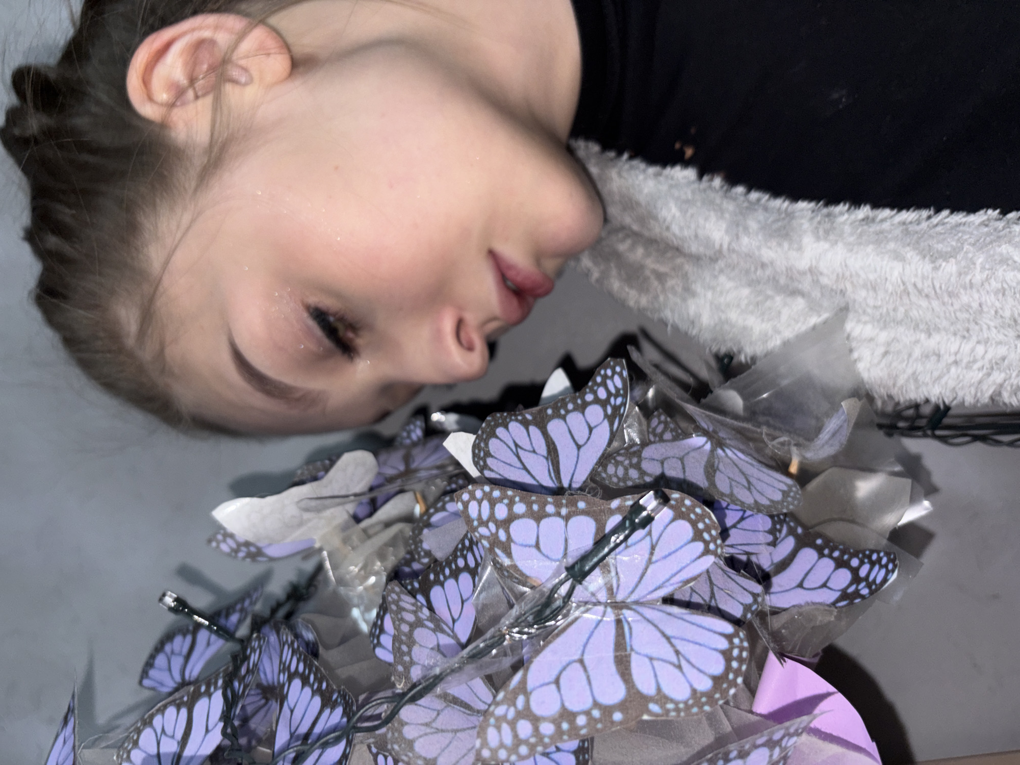 The image captures a close-up, angled portrait of a young person, likely female, from the chest up, in what appears to be an indoor setting in Chaikovsky, Russia. The person is lying on their side, with their head resting on a white, fluffy textile, possibly a towel or fleece blanket. They are wearing a dark-colored top, likely black. Their dark hair is pulled back, revealing an ear, and their eyes are closed or nearly closed, conveying a sense of peace or slumber. Subtle glitter or reflective specks are visible on their forehead and cheek.
In the foreground, a prominent cluster of artificial butterflies is arranged. These butterflies are primarily lavender or light purple with detailed black and white patterns, resembling monarch butterflies. Many of them are individually wrapped in clear plastic, suggesting they are part of a decorative display, possibly a bouquet or a string of lights, as a green wire is visible intertwined among them.
The scene is lit softly, suggesting an evening or nighttime setting, contributing to a calm and serene atmosphere. There is no visible text in the image. The overall impression is one of gentle repose amidst a whimsical, decorative arrangement.