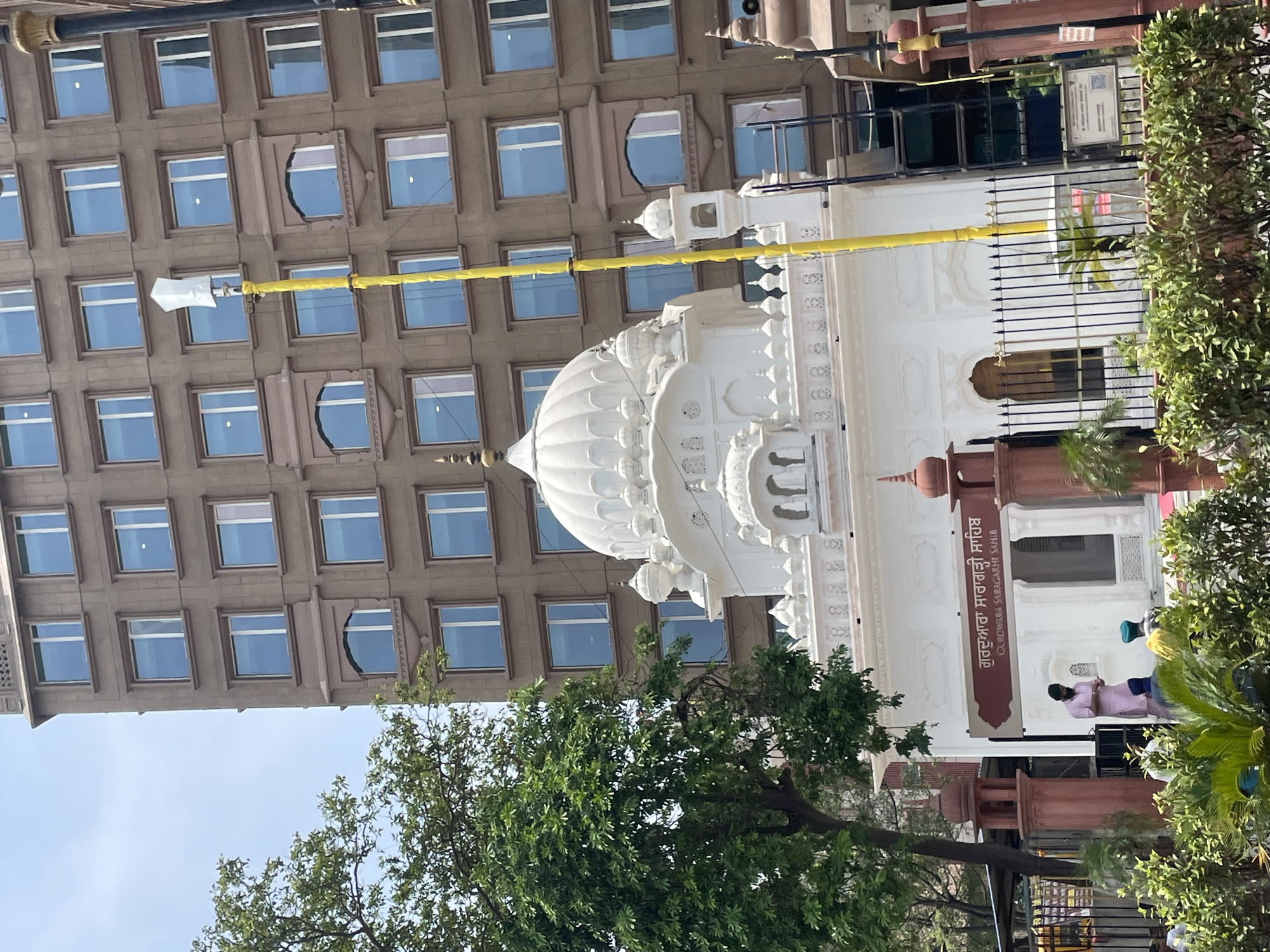 This is a medium-shot, eye-level photograph of the Gurdwara Saragarhi Sahib in Amritsar, India. The Gurdwara is a white marble building with a domed roof and ornate details, located in front of a tall, modern, brown brick building. A tall yellow pole, possibly a flag pole or antenna, stands to the right of the Gurdwara. The entrance to the Gurdwara has a sign with text in both Punjabi and English. Several people are visible, including a man in a pink kurta and a turban, and others wearing turbans. Green trees and bushes frame the foreground and left side of the image. The sky is visible and appears clear.