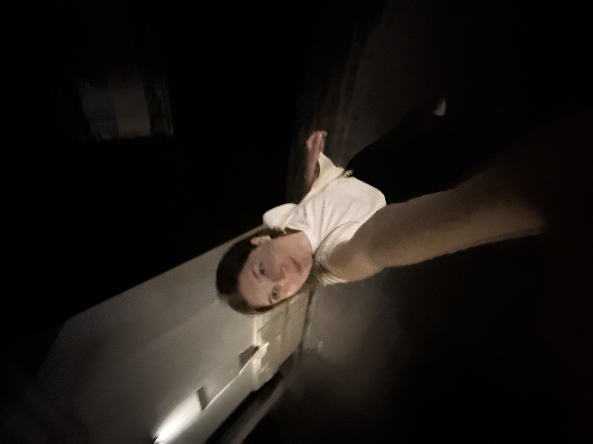 A low-angle, selfie-style photograph taken at night in Belgrade, Serbia. The subject is a person with light skin, dark hair, and wearing a white t-shirt, holding the camera at arm's length. The person is near the corner of a light-colored building or wall. The background is dark, with a possible outline of another building in the upper left corner.