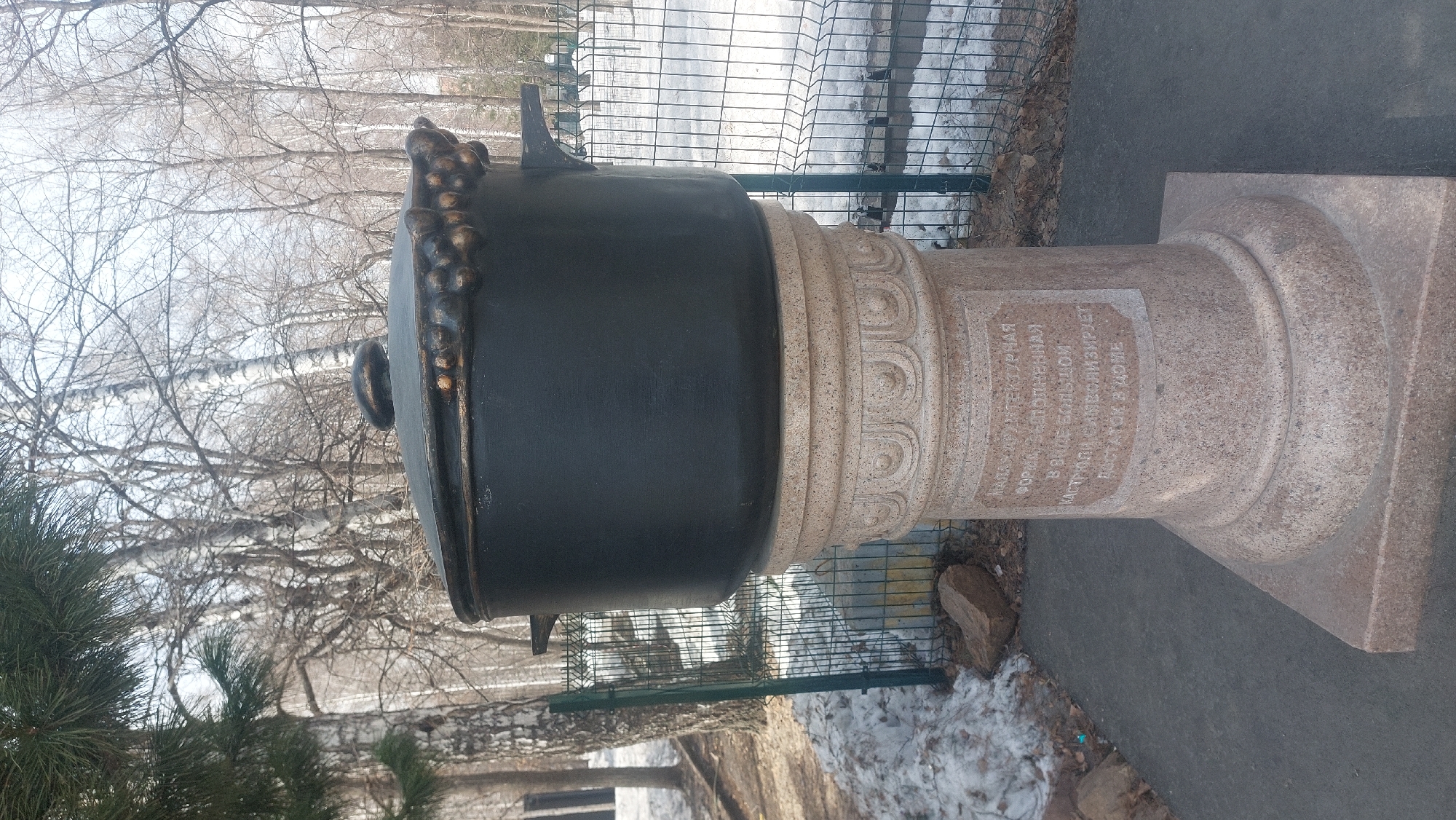 The image shows a monument in Novosibirsk, Russia. The monument consists of a large, black pot-shaped object resting atop a stone pedestal. The pedestal has decorative carvings and an inscription in Russian. The monument is located outdoors, near a sidewalk or paved area. In the background, there are trees without leaves, suggesting it is late fall or winter. A metal fence is also visible near the monument.