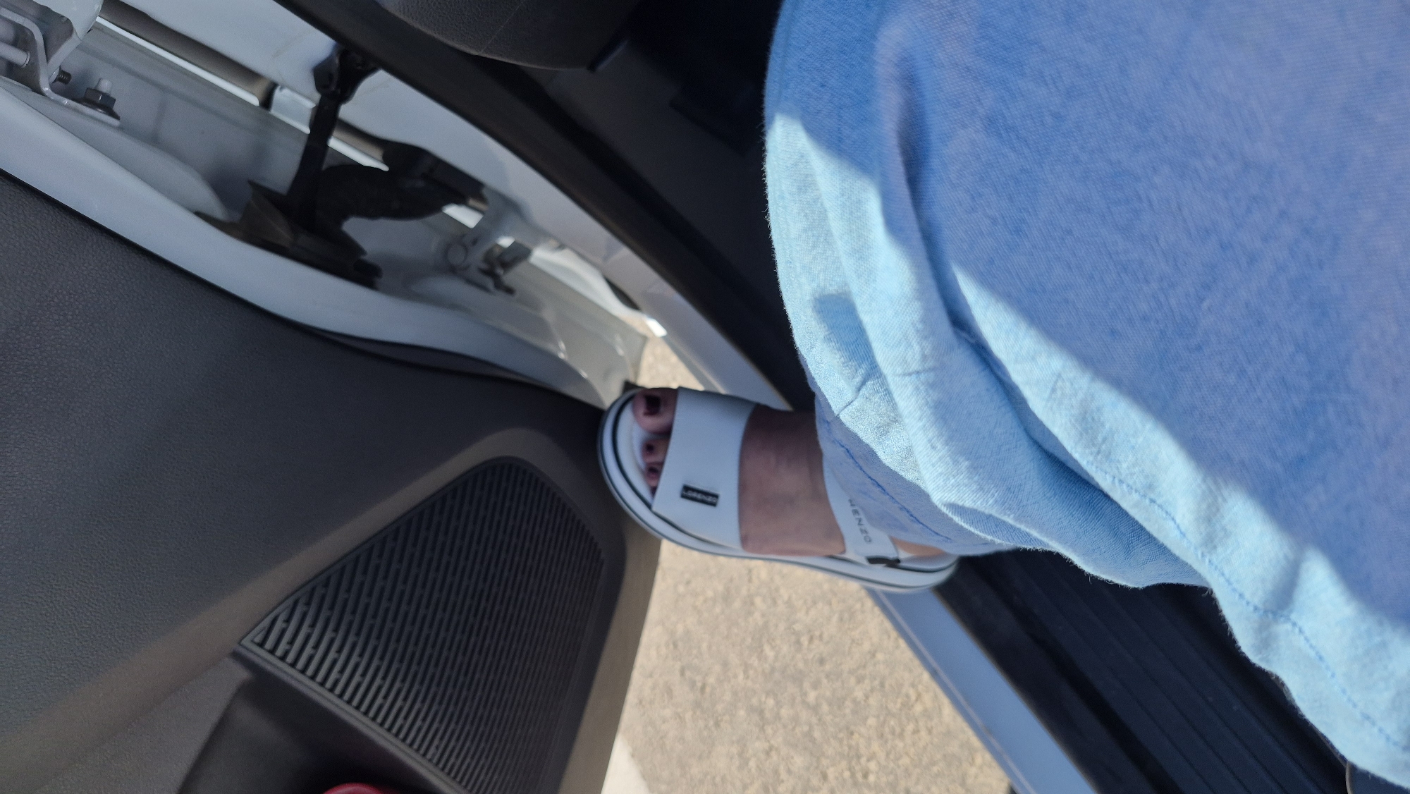 A first-person perspective shot shows a person's leg and foot inside a vehicle. The person is wearing light blue linen pants and a white sandal with a thick sole. The sandal has a single white strap across the toes and a black stripe along the edge of the sole. The foot is positioned on the car's door sill, with the car door ajar. The interior of the car is visible, including a dark gray door panel with a speaker grille. The exterior of the car is a bright white. The scene is likely in a parking area or on a street in Līvāni, Latvia, as indicated by the context.