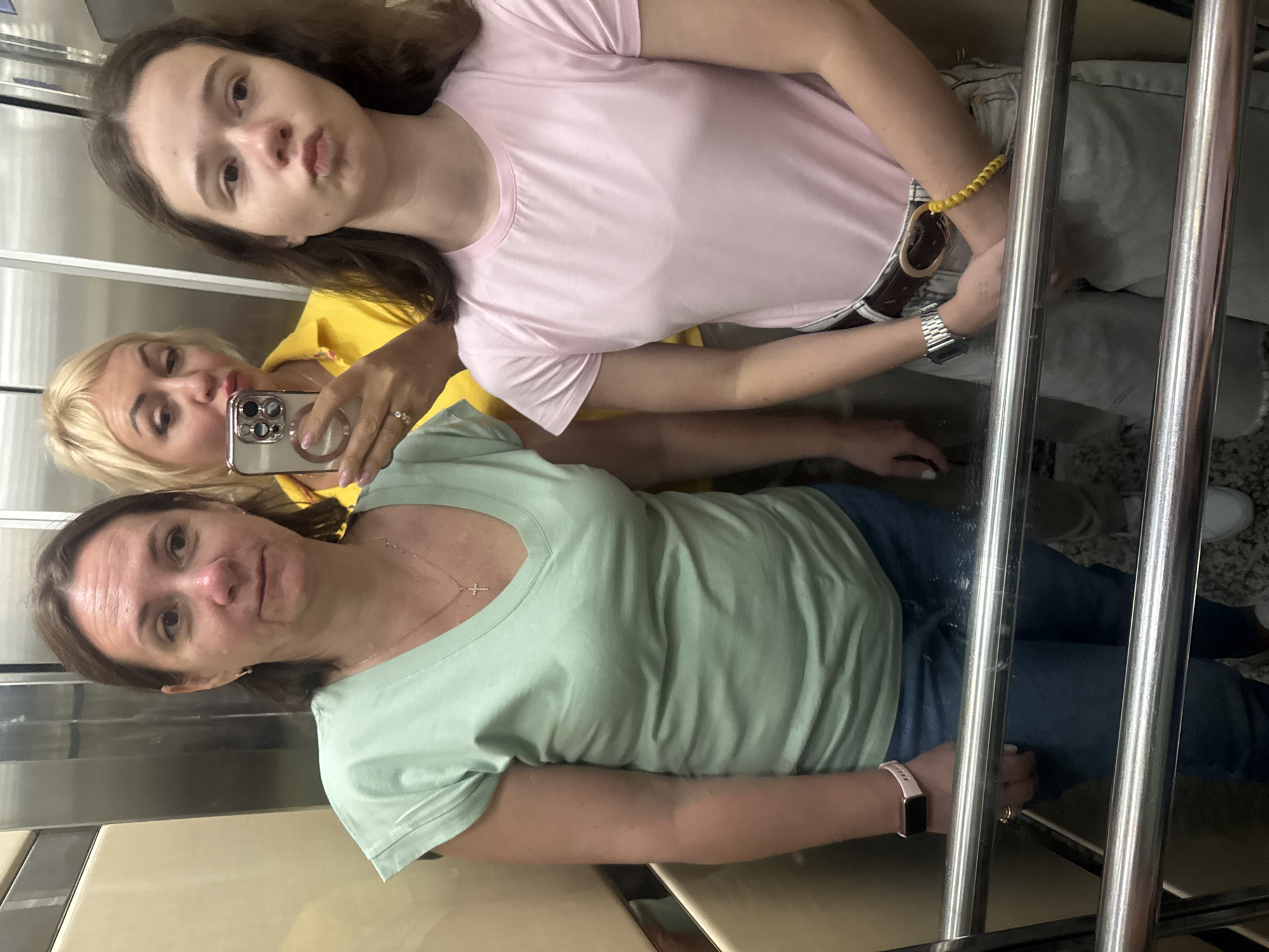Three women are taking a selfie in an elevator. The woman in the foreground is wearing a green shirt and jeans. The woman in the middle is wearing a yellow shirt. The woman in the background is wearing a pink shirt and gray pants.