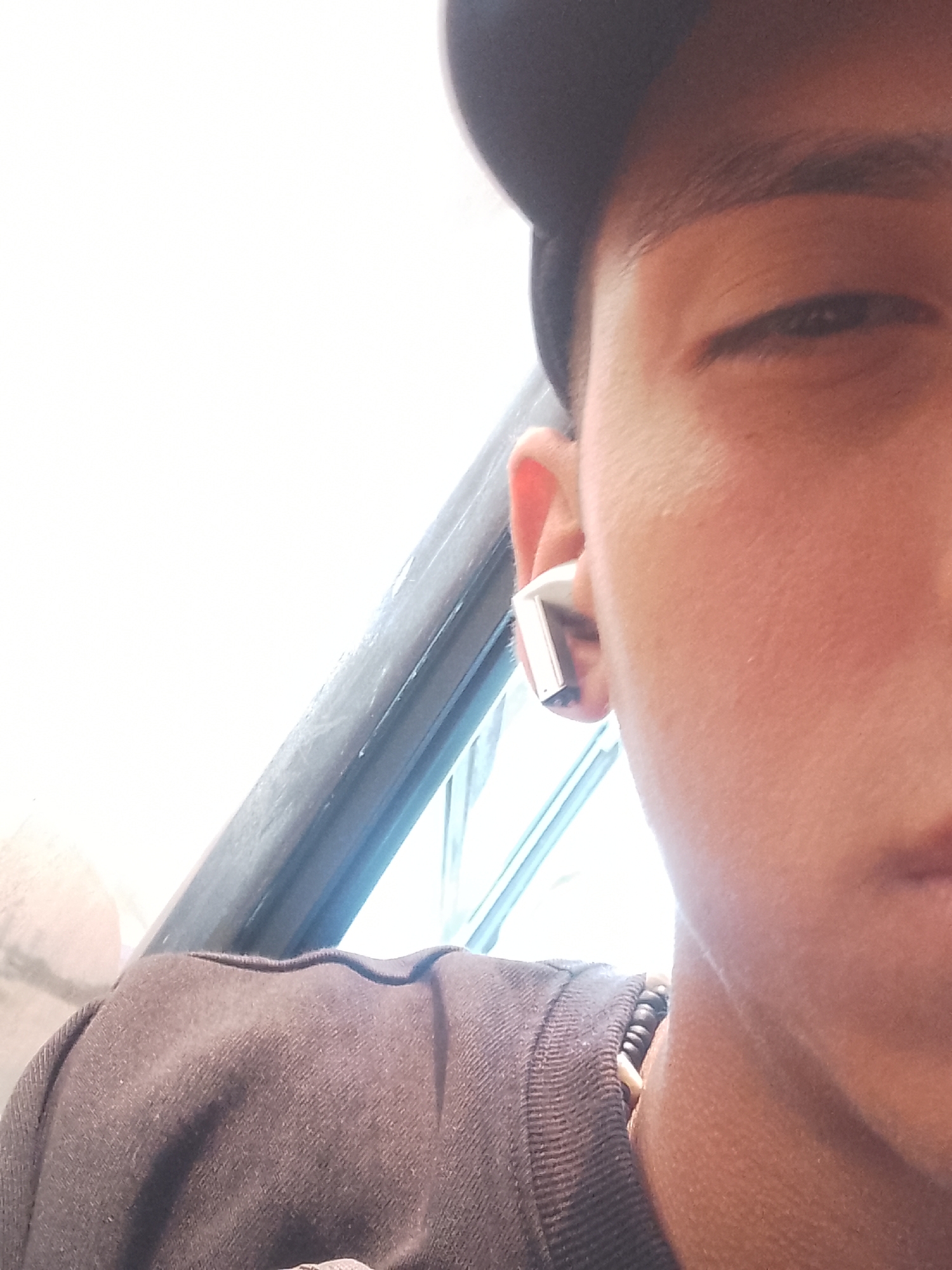 The image presents a close-up, angled portrait of a person, likely male, in Macho Muerto, Venezuela. The frame focuses on the right side of their face and extends down to their upper chest. The individual is wearing a dark-colored t-shirt, possibly black or dark brown, and a dark baseball cap, the brim of which is partially visible at the top of the head. A distinctive silver-colored earbud is worn in their right ear. Around their neck, a dark, beaded necklace or similar accessory is partially visible.
The person has a neutral expression, with their right eye looking towards the camera. The background on the left side of the image is extremely bright and overexposed, indicating strong natural light, suggesting it is daytime and likely sunny. A dark, possibly metallic, bar or railing runs diagonally through the mid-ground behind the person, creating a visual separation between the bright sky-like area and a slightly darker, indistinct area below it. This element could be part of a vehicle, a building structure, or an outdoor railing. There is no visible text in the image.