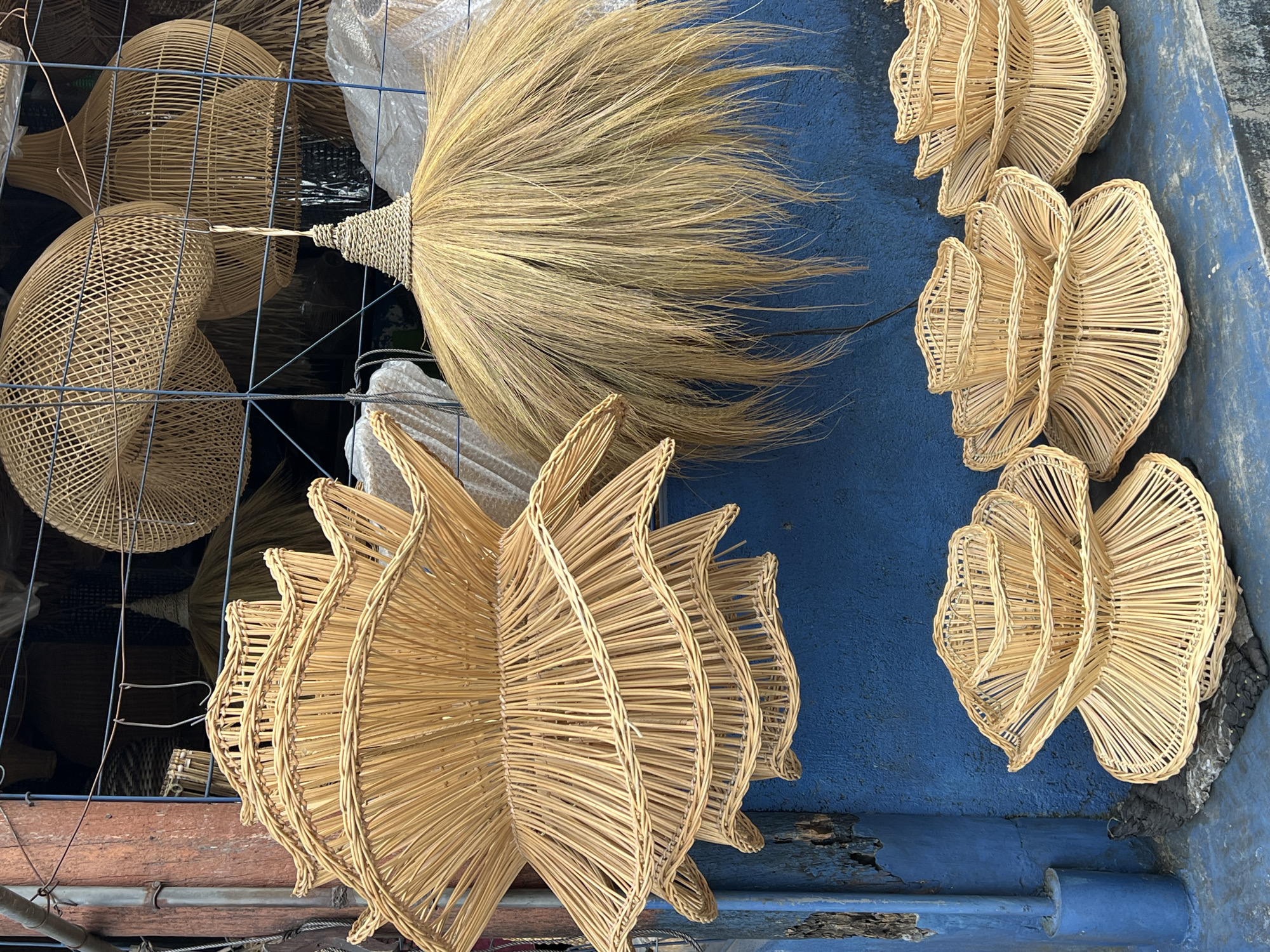 The image shows a collection of handcrafted items displayed on a blue surface. On the left, there are several woven baskets with a dome-like shape, some behind a wire mesh. A large, dried grass broom with a woven handle is positioned centrally. To the right, there are numerous decorative items made of woven material, formed into repeating petal-like shapes. The setting is likely a marketplace or shop in the Ubud District, Indonesia, indicated by the style of the artisanal crafts. There are no people visible. The environment suggests an outdoor setting or a shopfront.