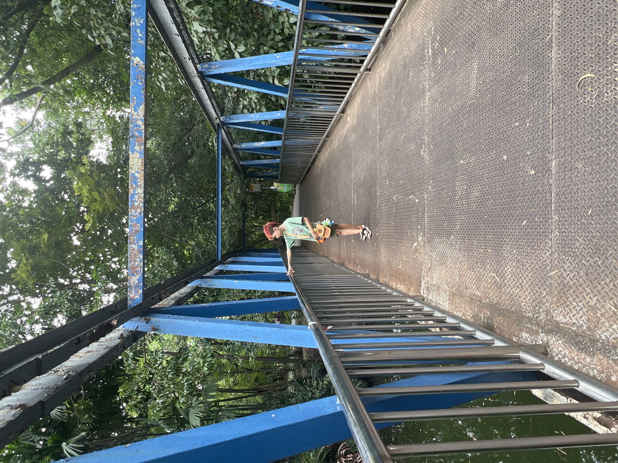 The image shows a young person with red hair, wearing a green t-shirt, brown shorts, and sandals, standing on a metal pedestrian bridge in Bangkok, Thailand. The bridge has bright blue painted metal supports and railing, with stainless steel vertical bars along the sides. The bridge deck is made of diamond plate metal. The person is reaching out to touch the railing and looking to the left. In the background, there is lush green foliage and trees, indicating a park or natural area. A body of water with some floating leaves is visible to the left of the bridge. The overall scene suggests a relaxed outdoor setting within the city.