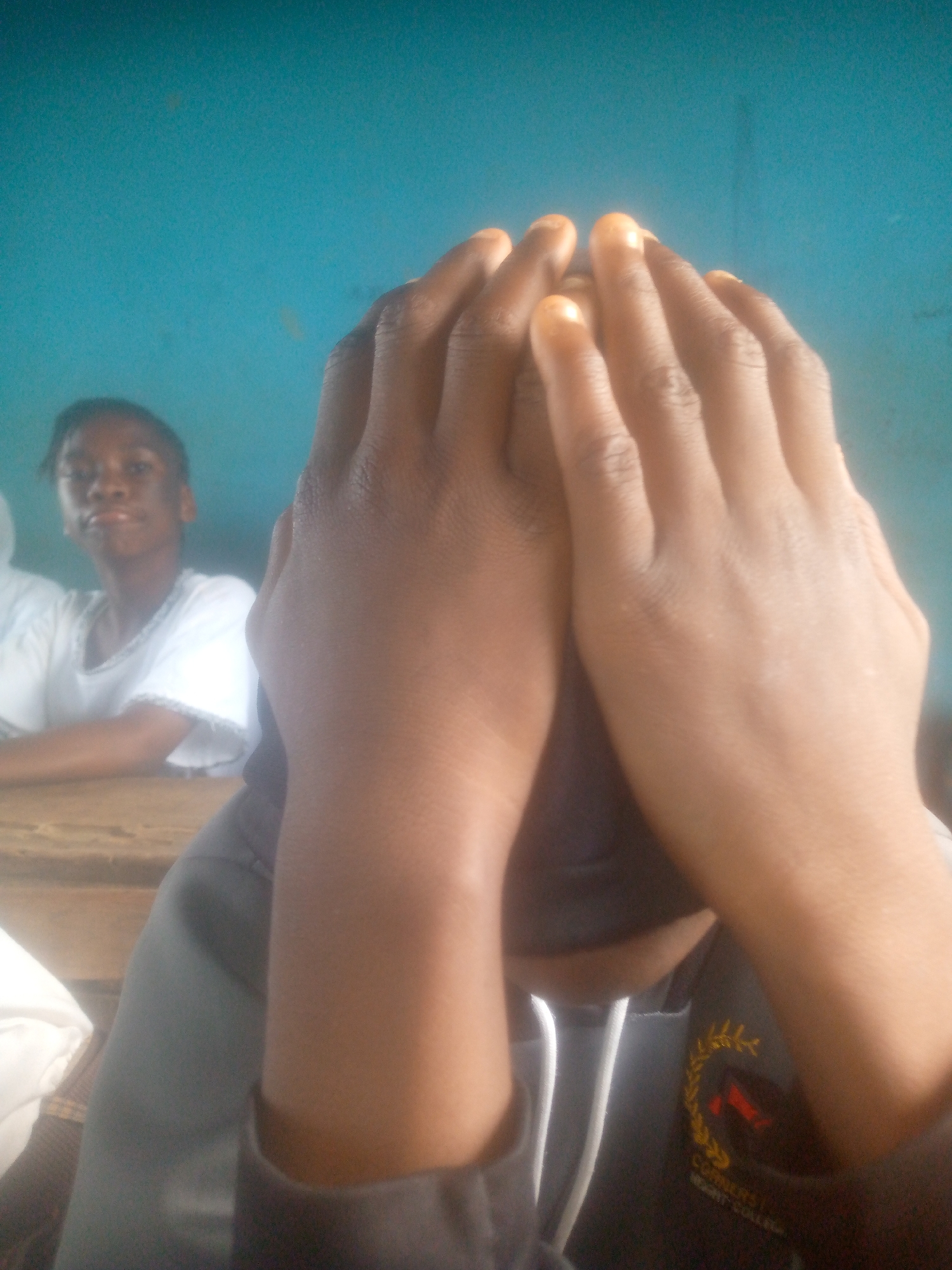 The photo shows a close-up of a person sitting at a table, with their hands covering their face. A young woman in a white shirt is seated at the same table in the background. The setting appears to be indoors with a blue wall. A school logo is visible on the person's clothes. The photo was taken in Ikorodu, Nigeria.