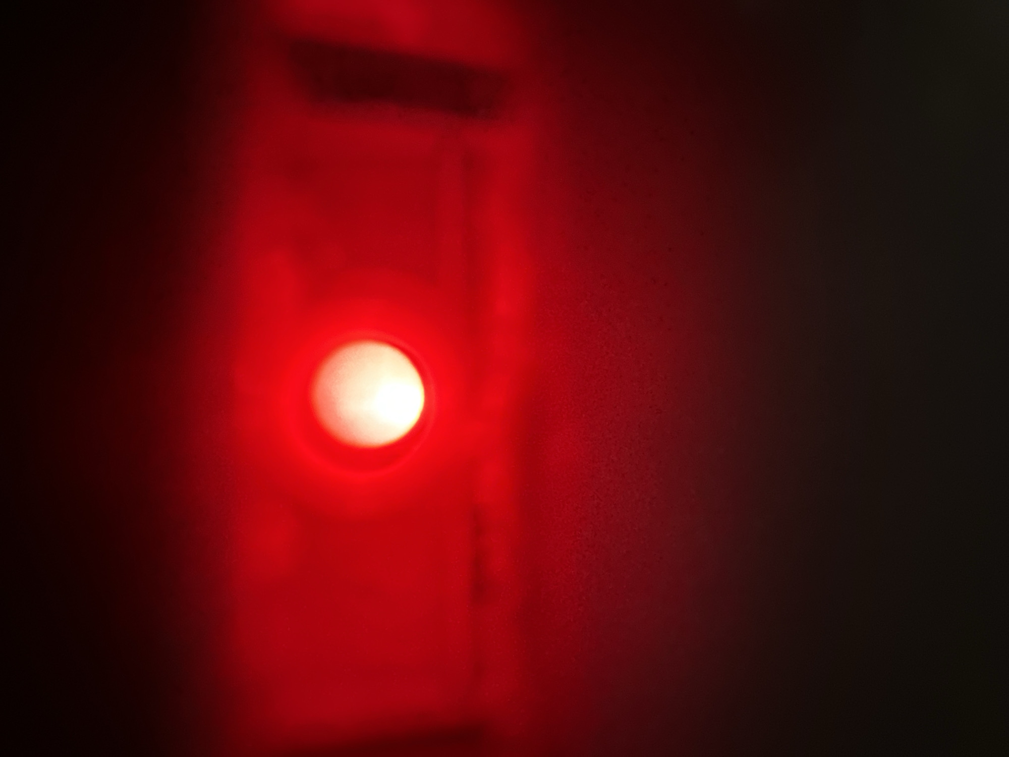 The image is a close-up, low-light photograph with a red hue. The primary subject is a bright, circular light source, likely an LED, situated slightly left of center. This light emanates a strong red glow that saturates the left side of the frame, fading into darkness towards the right. The light source appears to be embedded within a dark, rectangular recess or panel, with some indistinct lines or edges visible within this area. The surface surrounding the light appears to be textured, with a grainy quality throughout the image, particularly noticeable in the red-lit areas and the darker right side. The overall impression is one of a dimly lit environment, possibly an electronic device or a dark room with a single illuminated indicator. There is no discernible text visible in the image. The setting is not identifiable beyond its general appearance as a man-made structure or device.